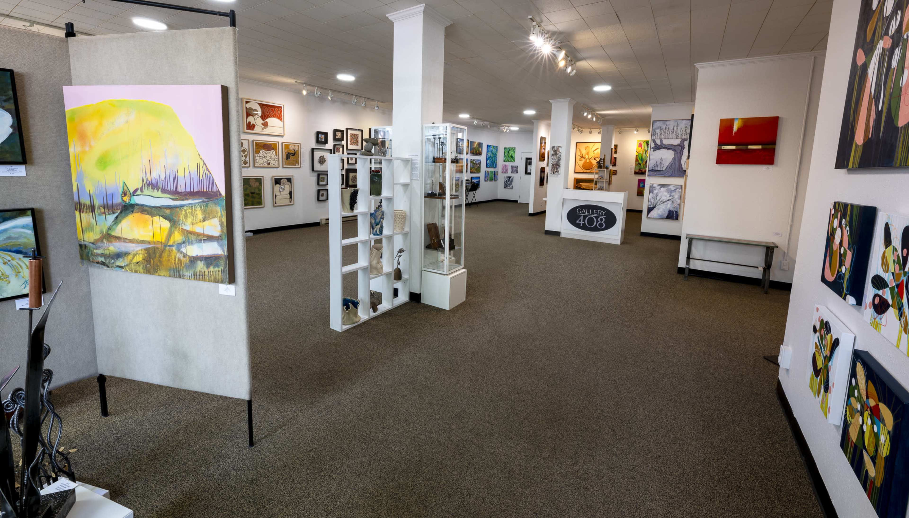 The image shows an interior view of an art gallery featuring various framed artworks displayed on the walls and stands.