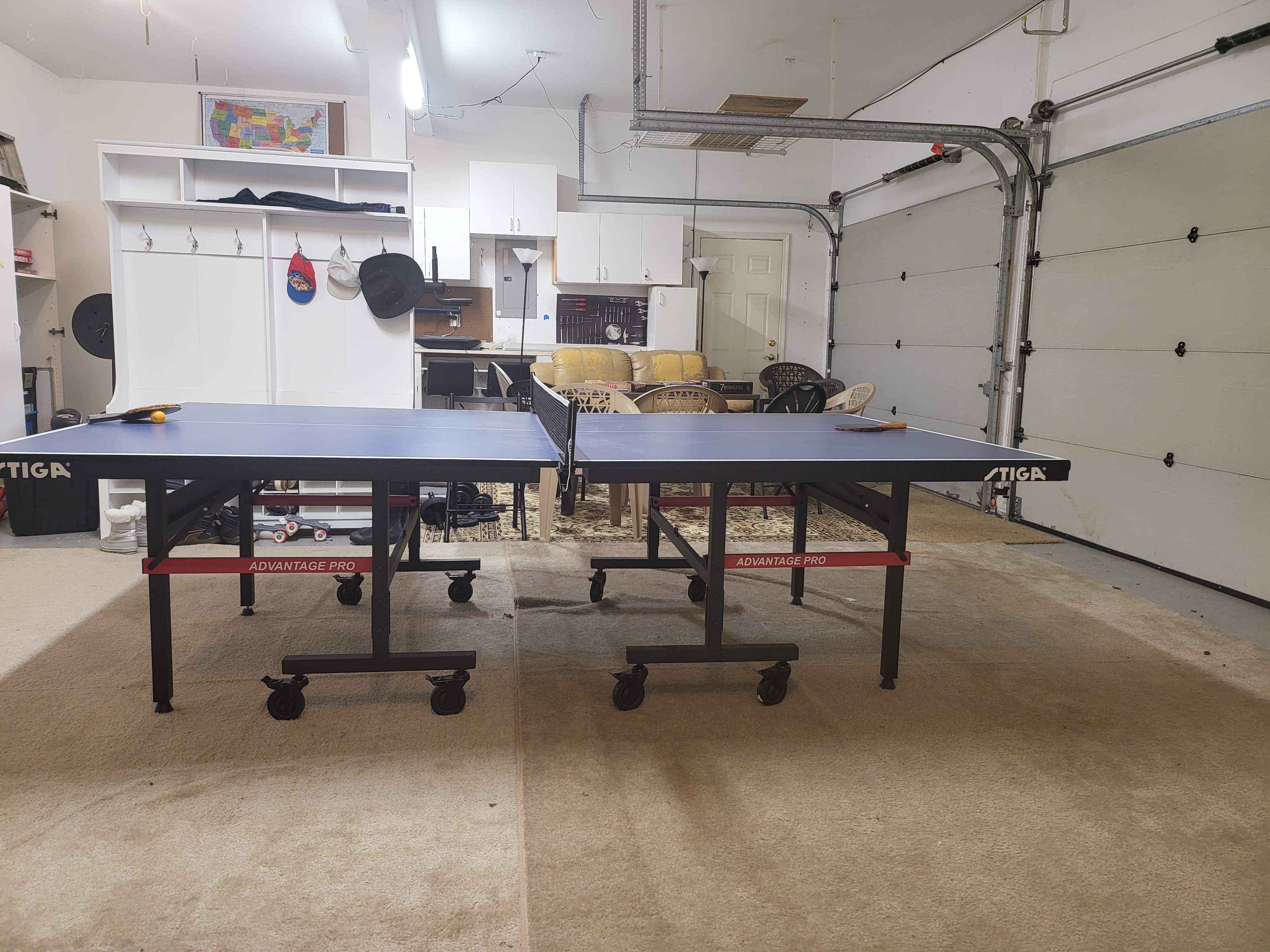 Two table tennis tables are set up in a garage, surrounded by various furniture and storage items.