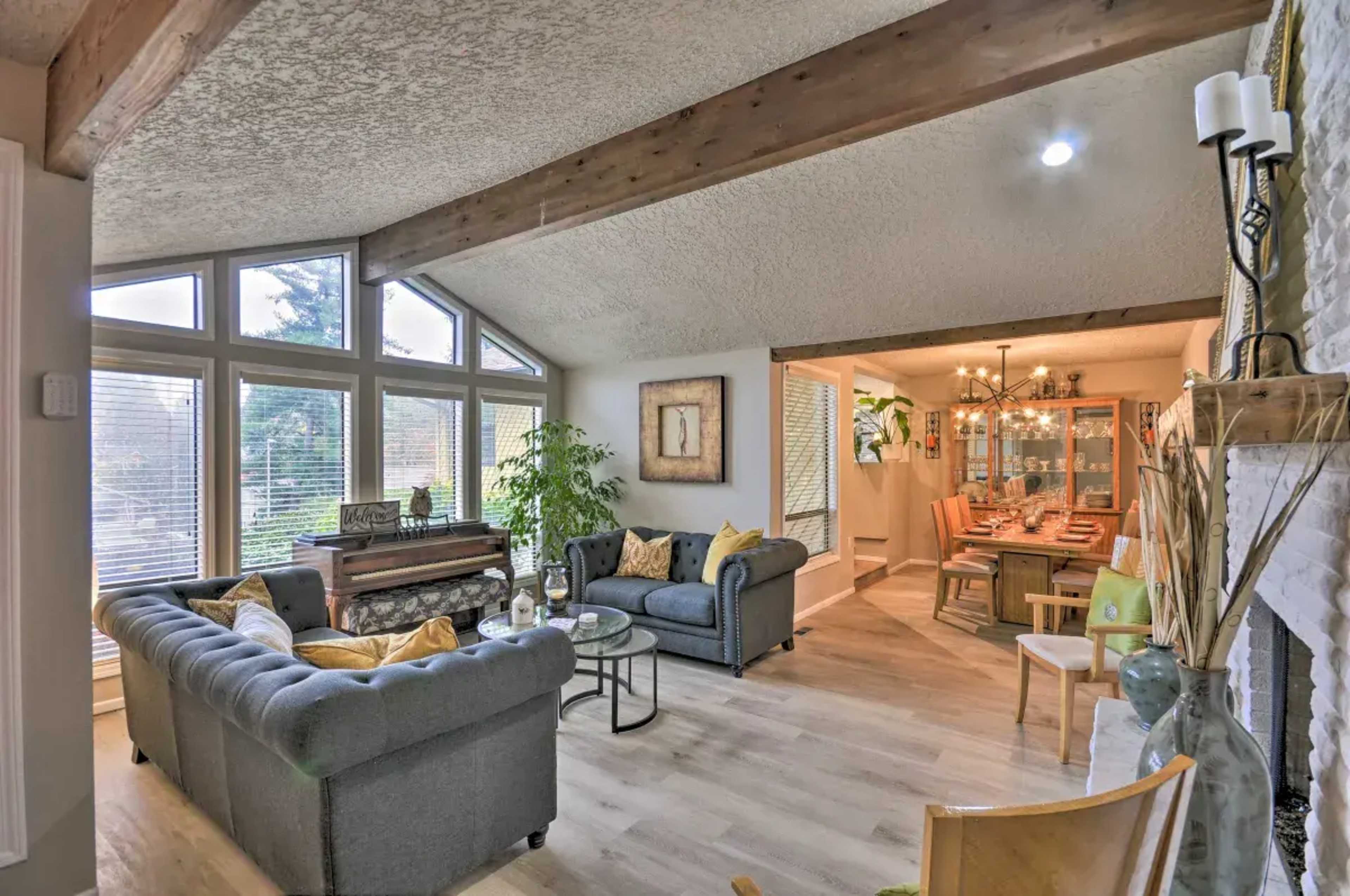 A living room with a piano, two sofas, and large windows allowing natural light, leading into a dining area visible in the background.