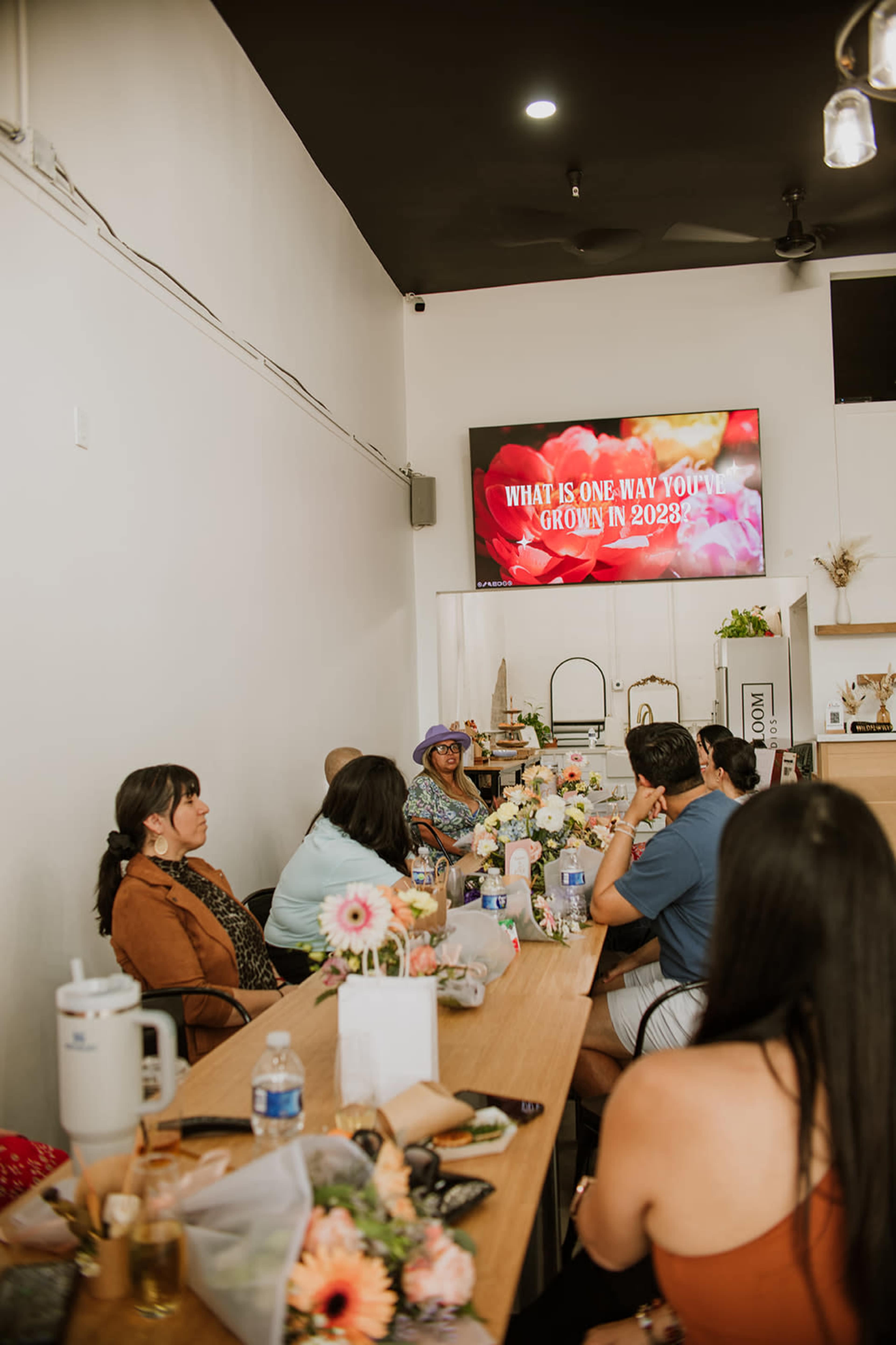 A group of people sits around a long table in a bright room, with a screen displaying the question, "What is one way you want to grow in 2023?" in the background.