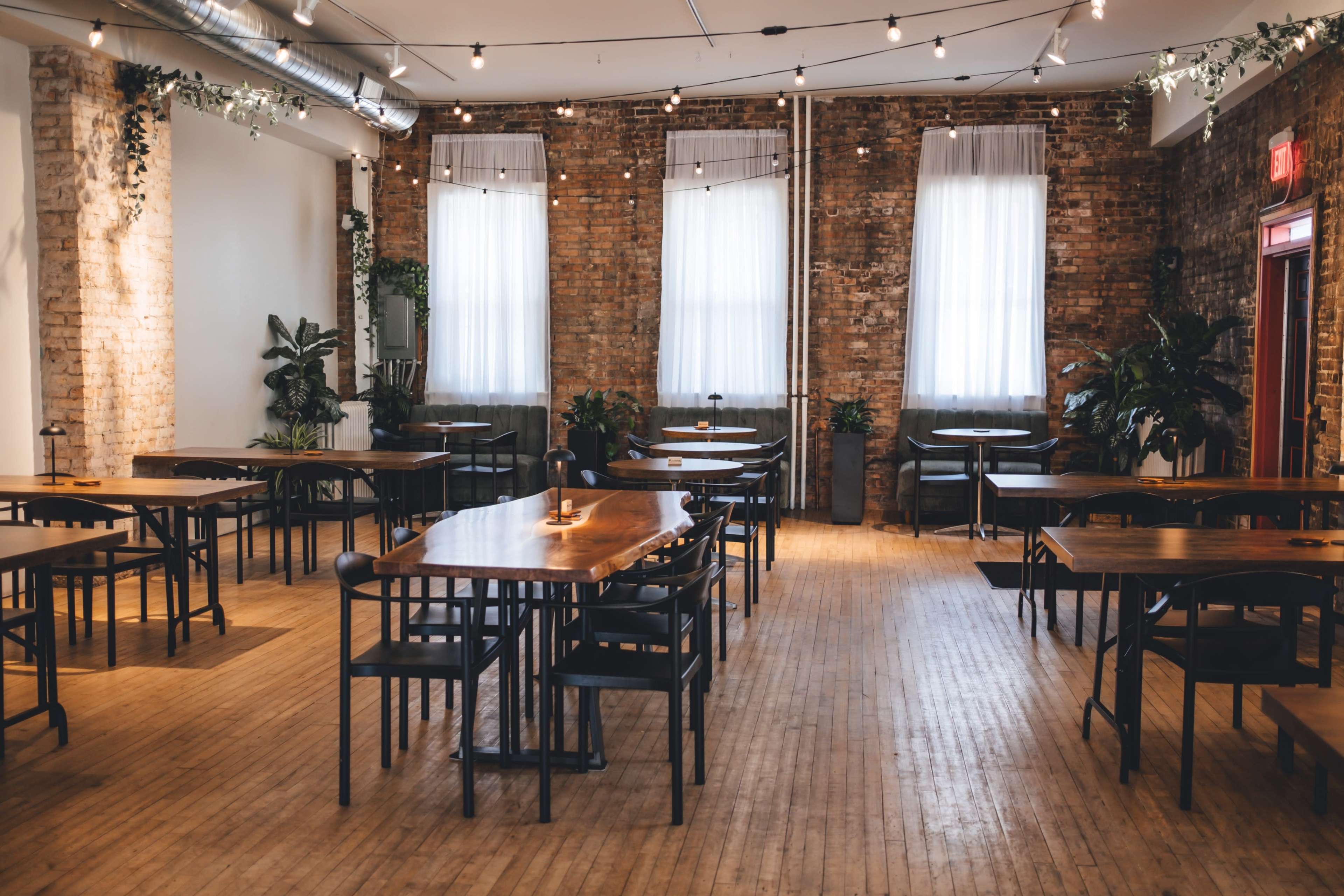 A spacious indoor dining area features several wooden tables surrounded by dark chairs, with large windows letting in natural light and numerous indoor plants.