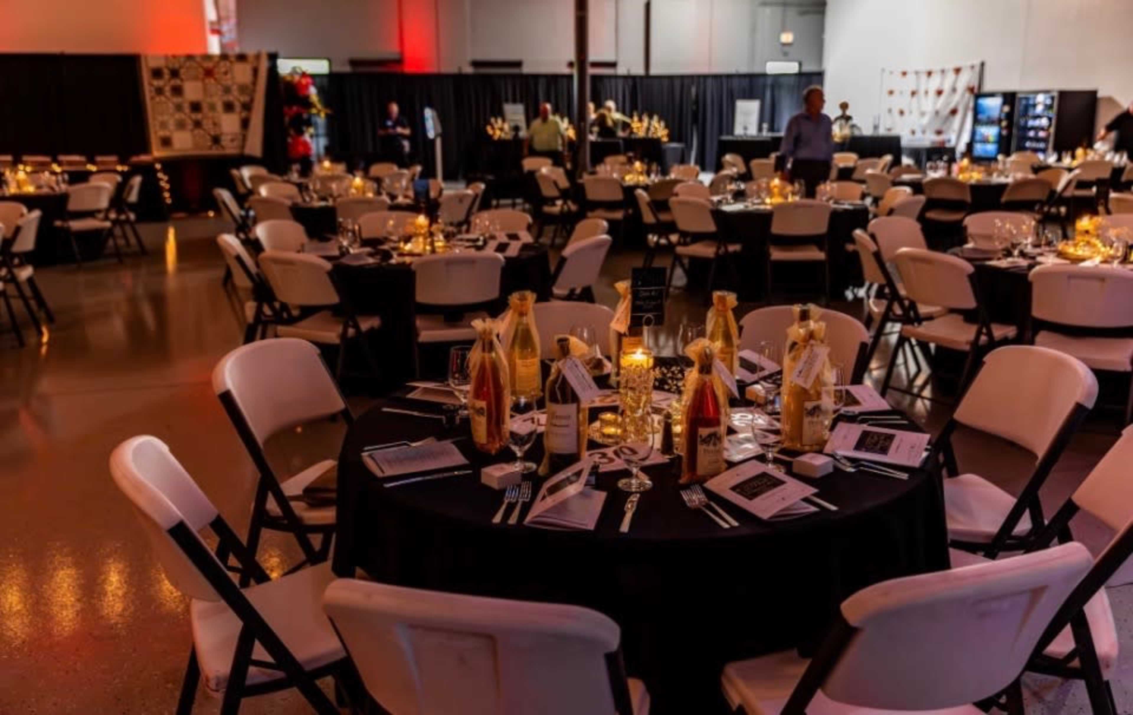 The image shows a banquet hall set with round tables covered in black tablecloths, each adorned with elegant place settings and decorative bottles, while people socialize in the background.