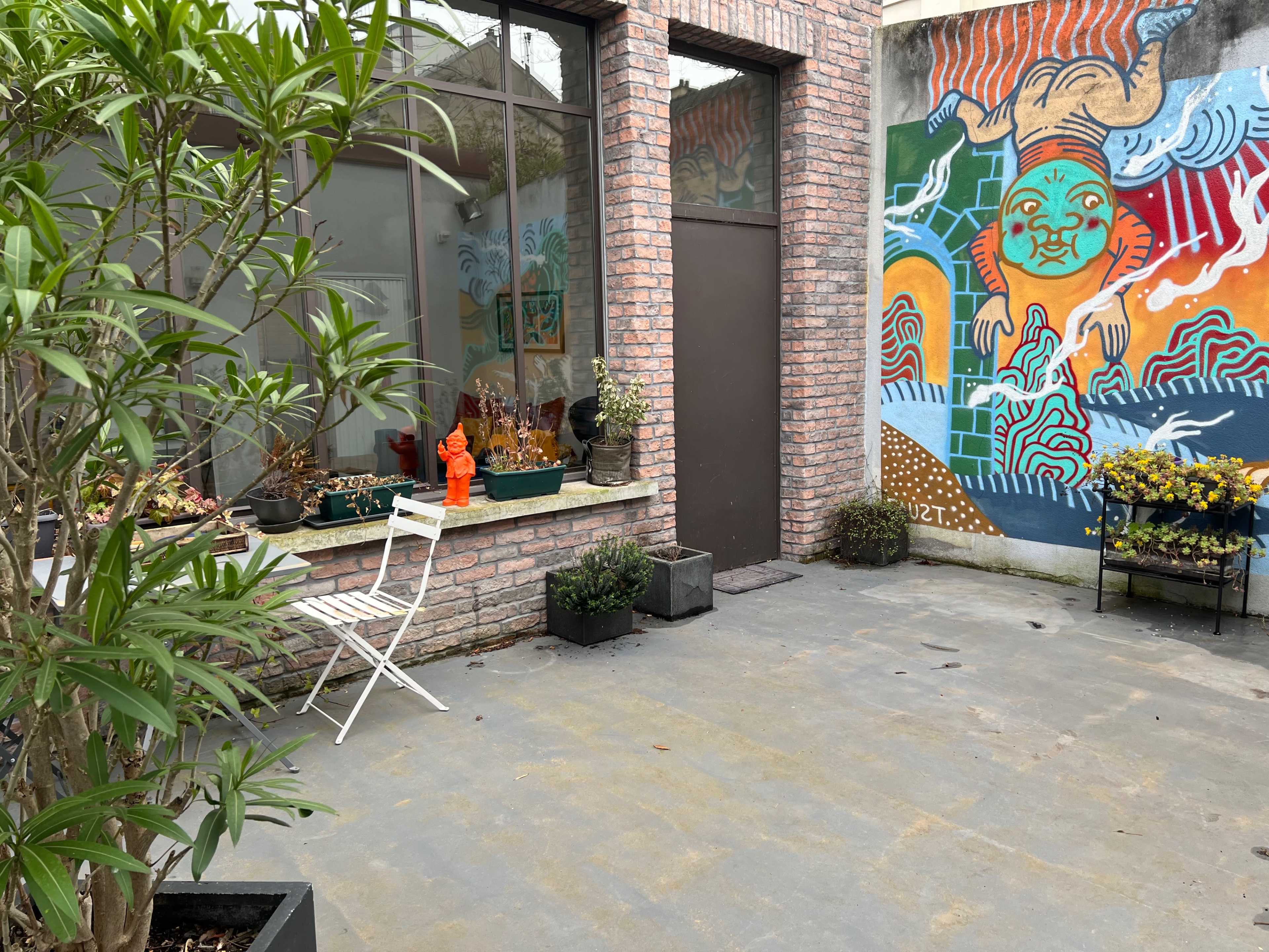 A small outdoor space featuring potted plants, a white metal chair, and a colorful mural on a brick wall.