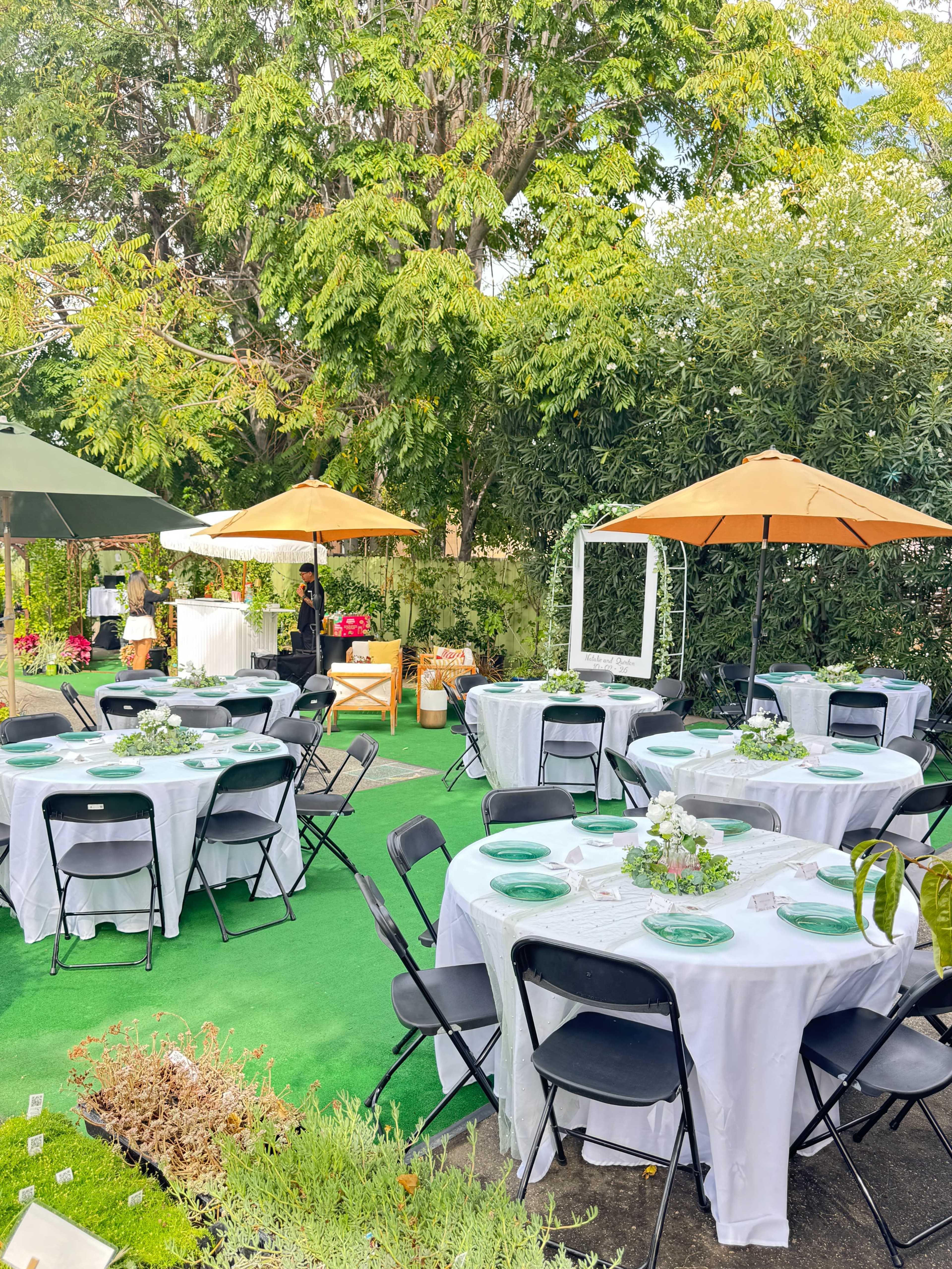 The image shows an outdoor dining setup with round tables covered in white tablecloths, black chairs, and orange umbrellas in a landscaped garden.