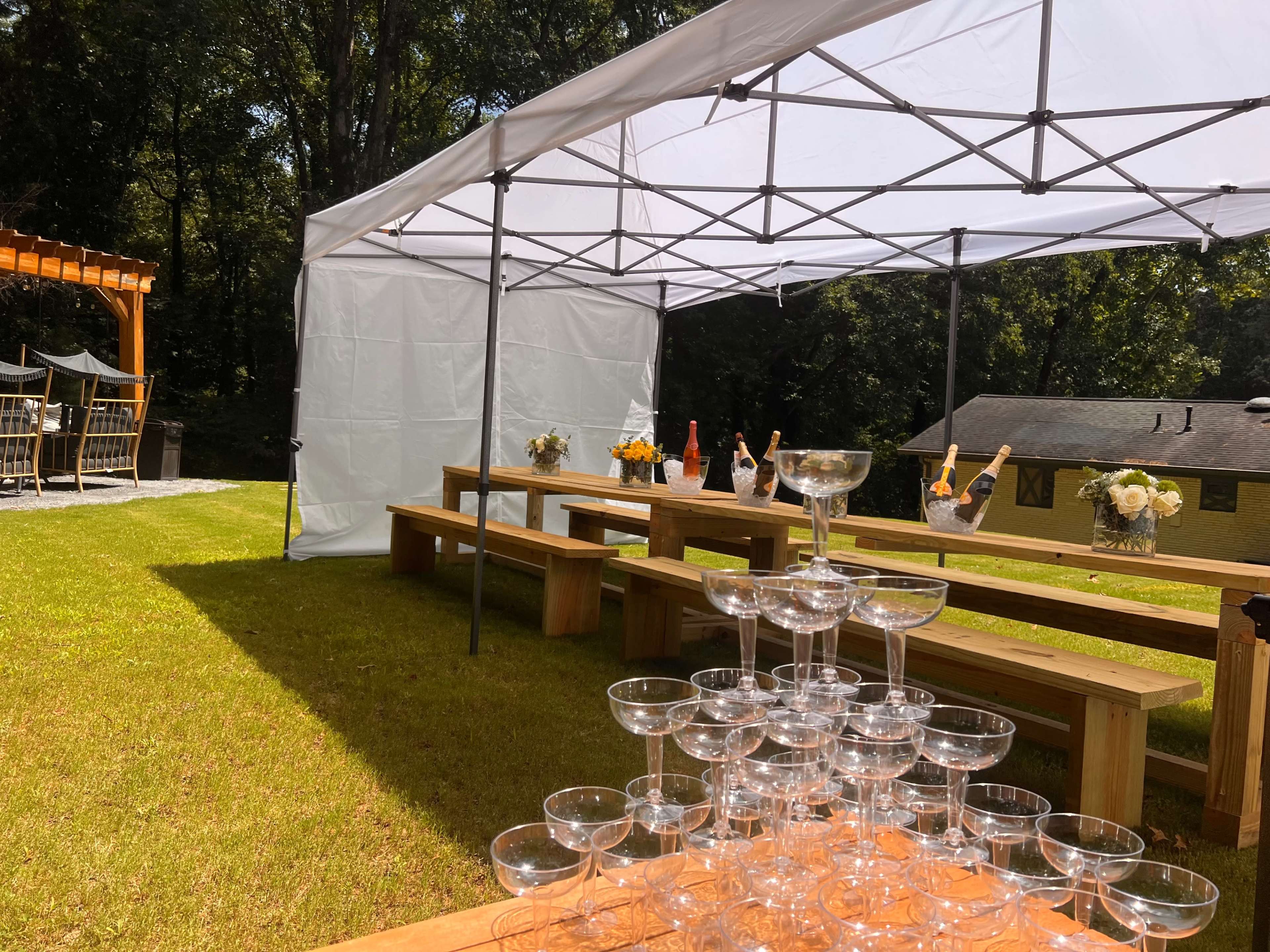 A white canopy is set up outdoors over wooden benches, with a display of champagne glasses and bottles arranged on a table in the background.
