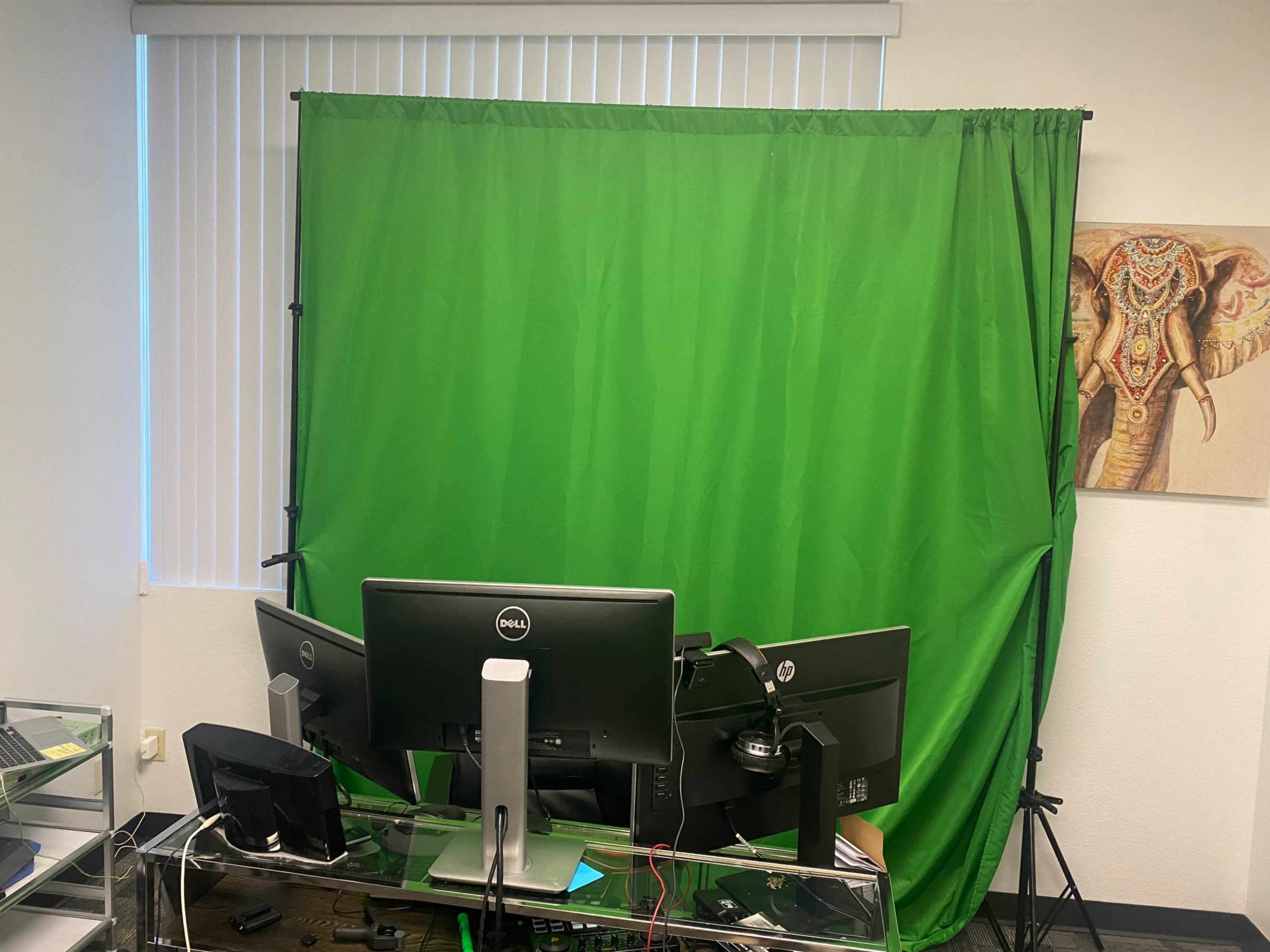 The image shows a workspace featuring a green screen setup behind dual computer monitors, with a decorative elephant painting on the wall.