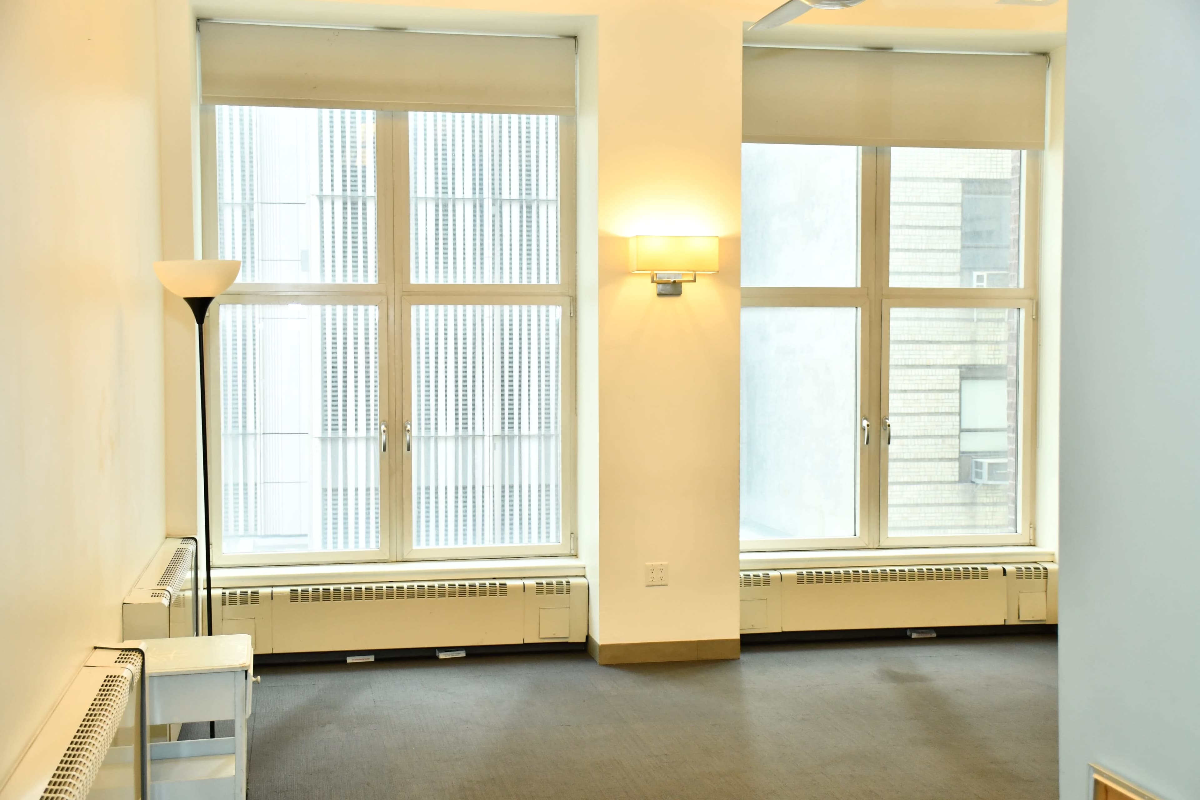 The image shows a room with large windows on two walls, minimal furniture including a lamp and a small table, and a carpeted floor.