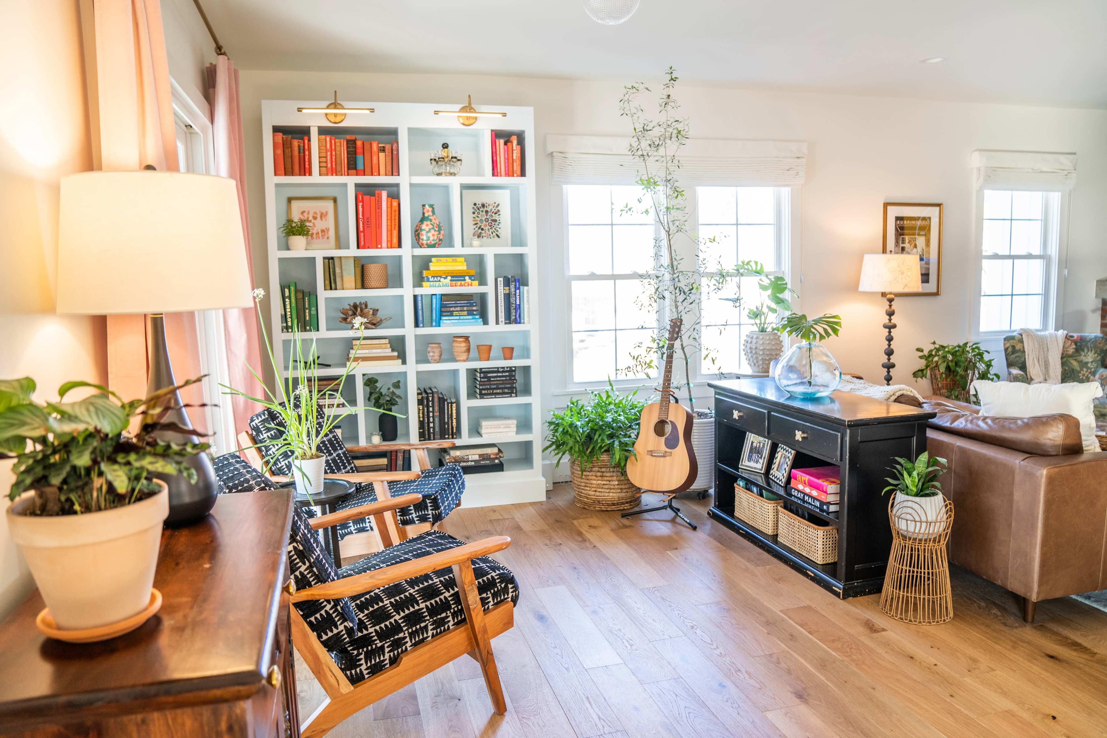 A bright living room with a bookshelf filled with books, a guitar leaning against the wall, and several plants in decorative pots.