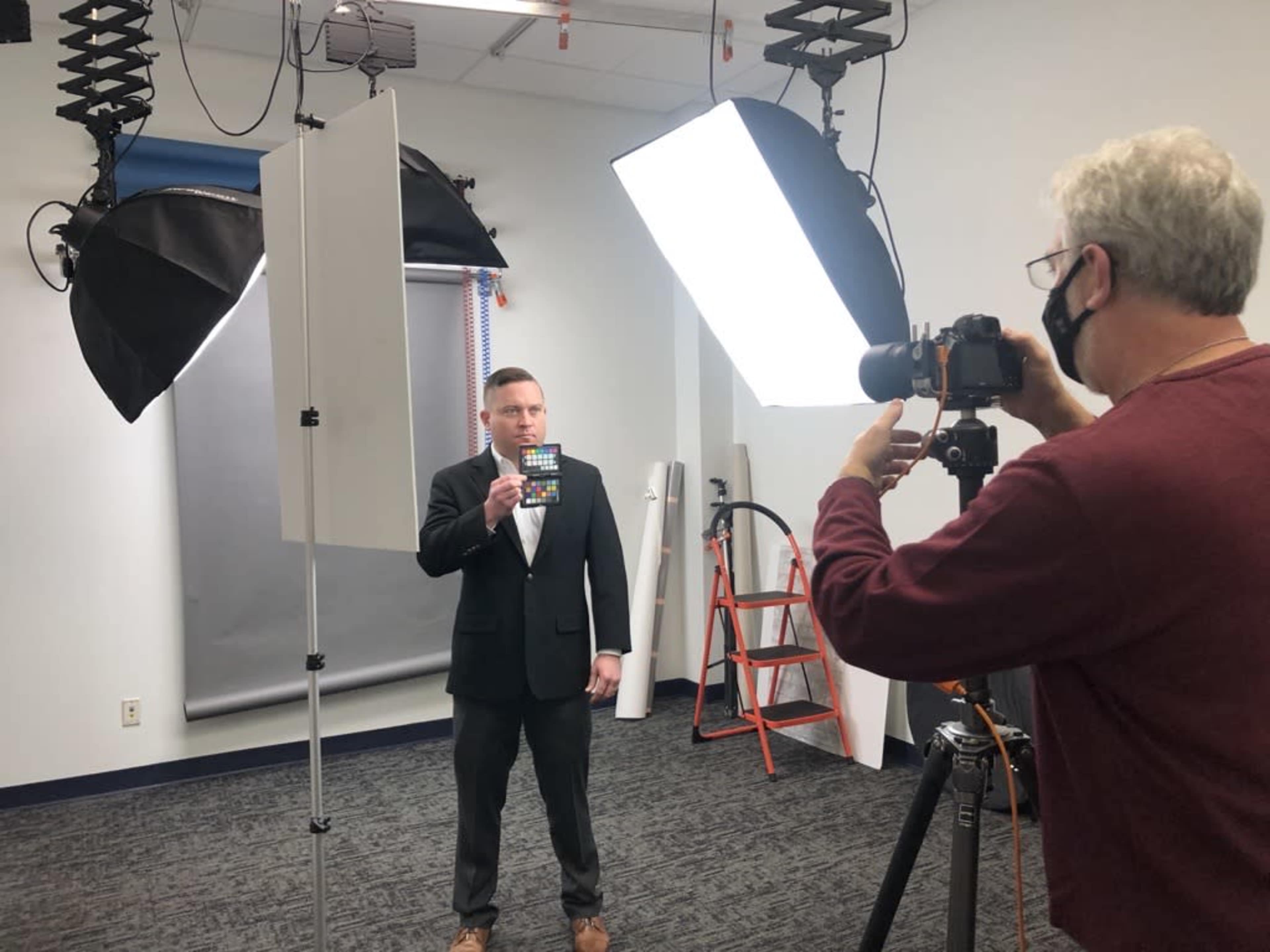 A man in a suit holds a slate while being photographed by another man in a studio setting with lighting equipment.