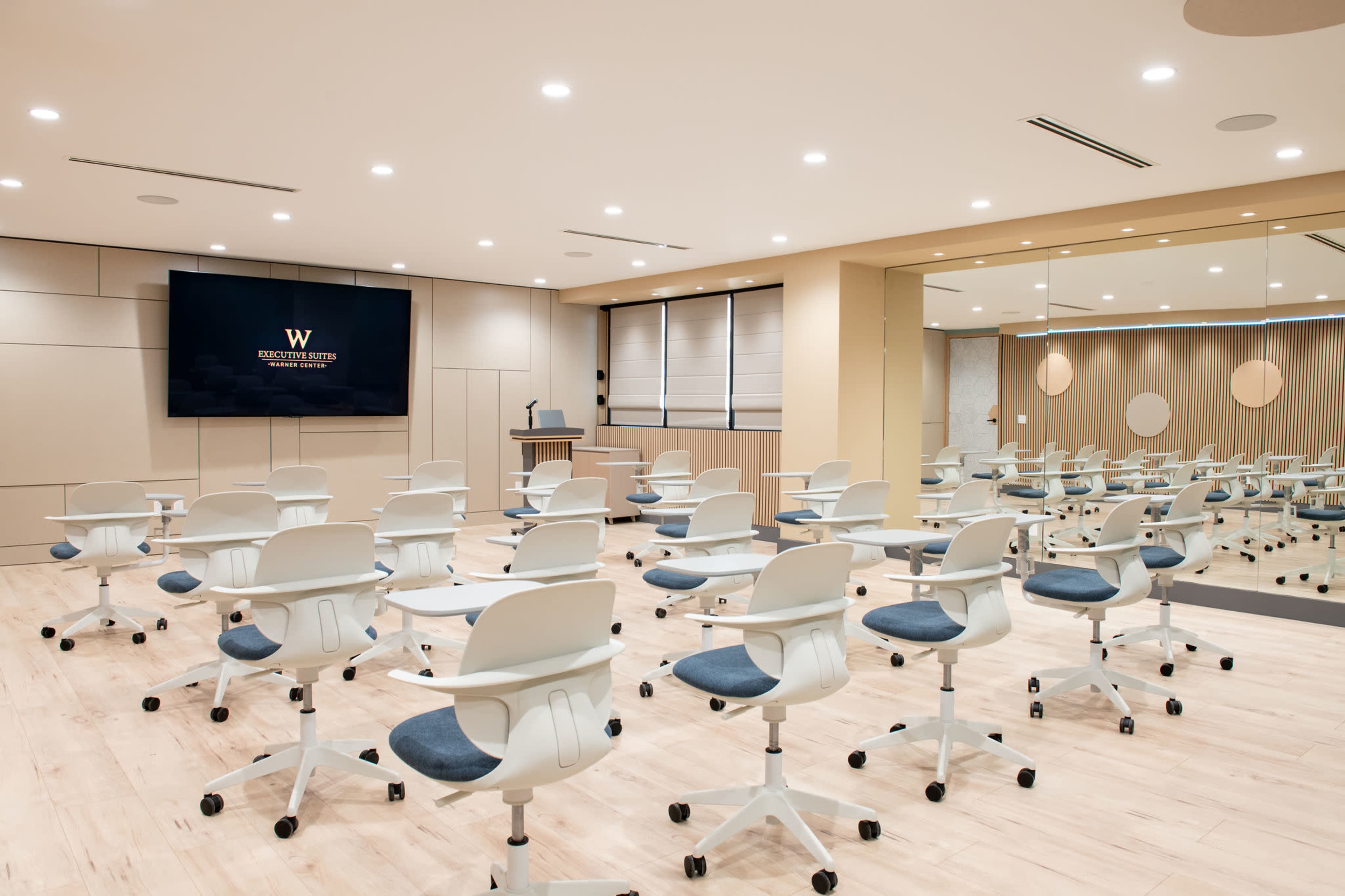 The image shows a modern classroom setting with rows of light-colored rolling chairs arranged in front of a large screen and a podium.