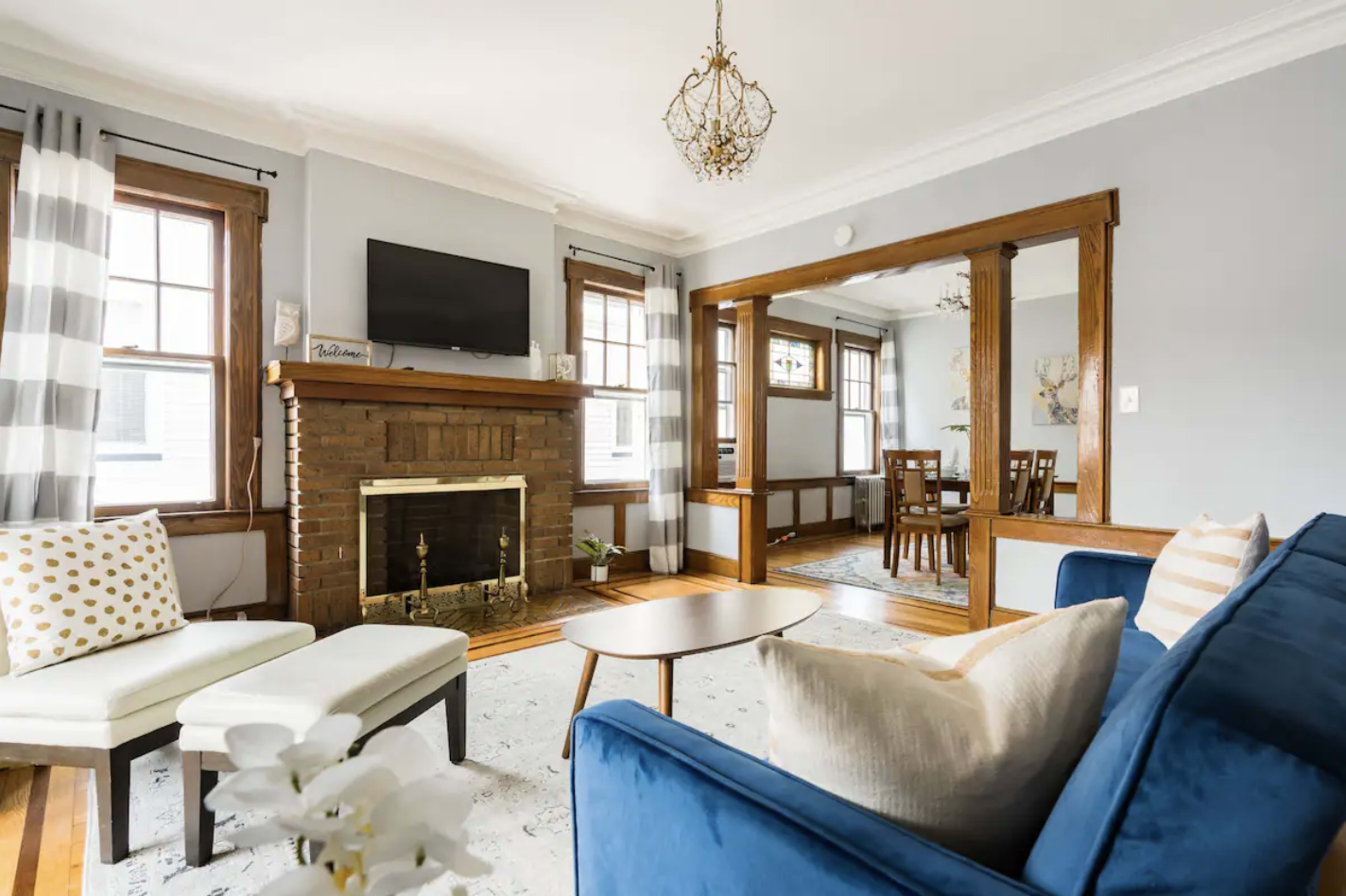 The living room features a brick fireplace with a television above it, a blue sofa, a patterned rug, and a dining area visible through an open archway.