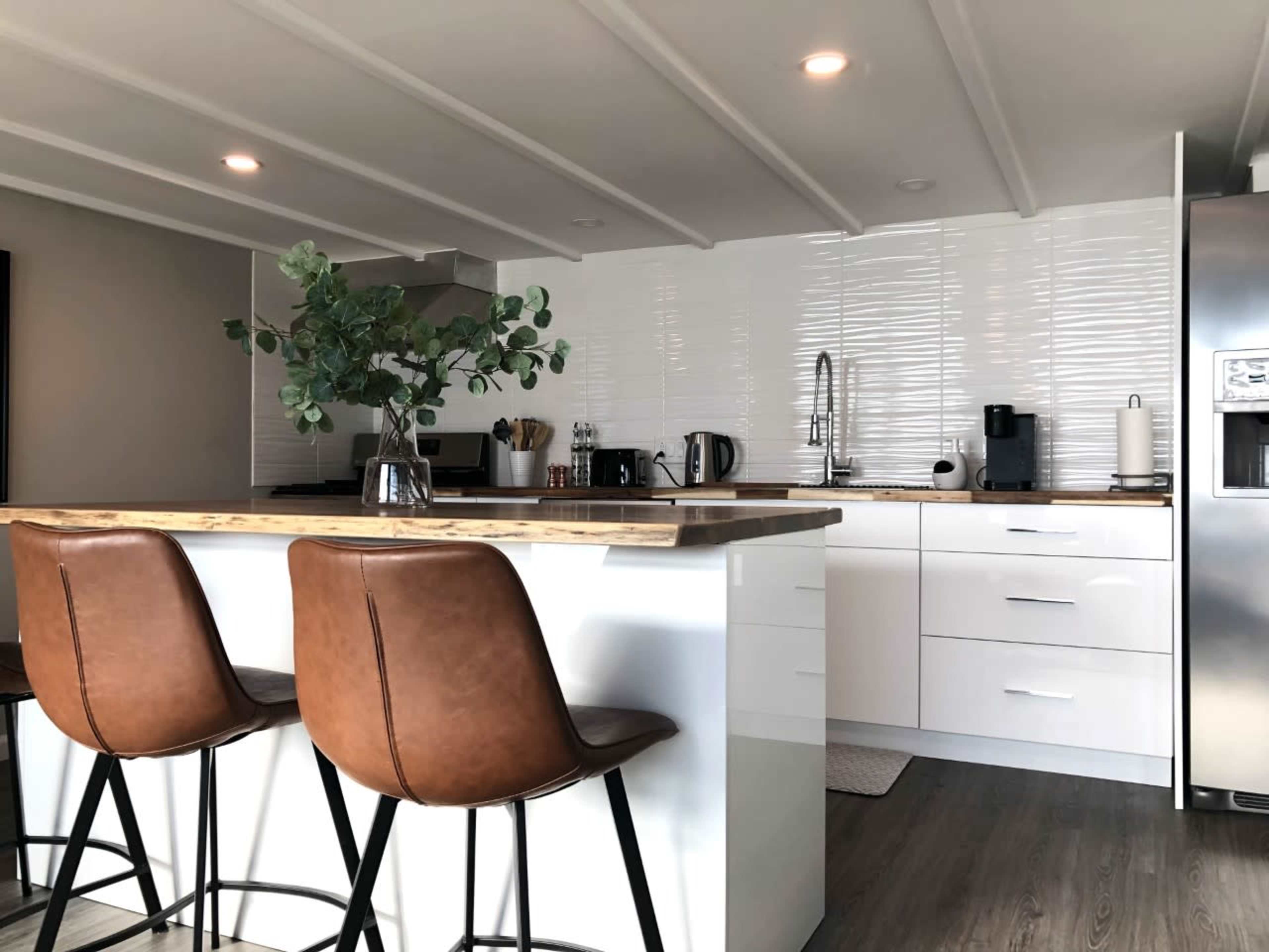 A modern kitchen features a wooden countertop island with brown bar stools, white cabinetry, and stainless steel appliances.