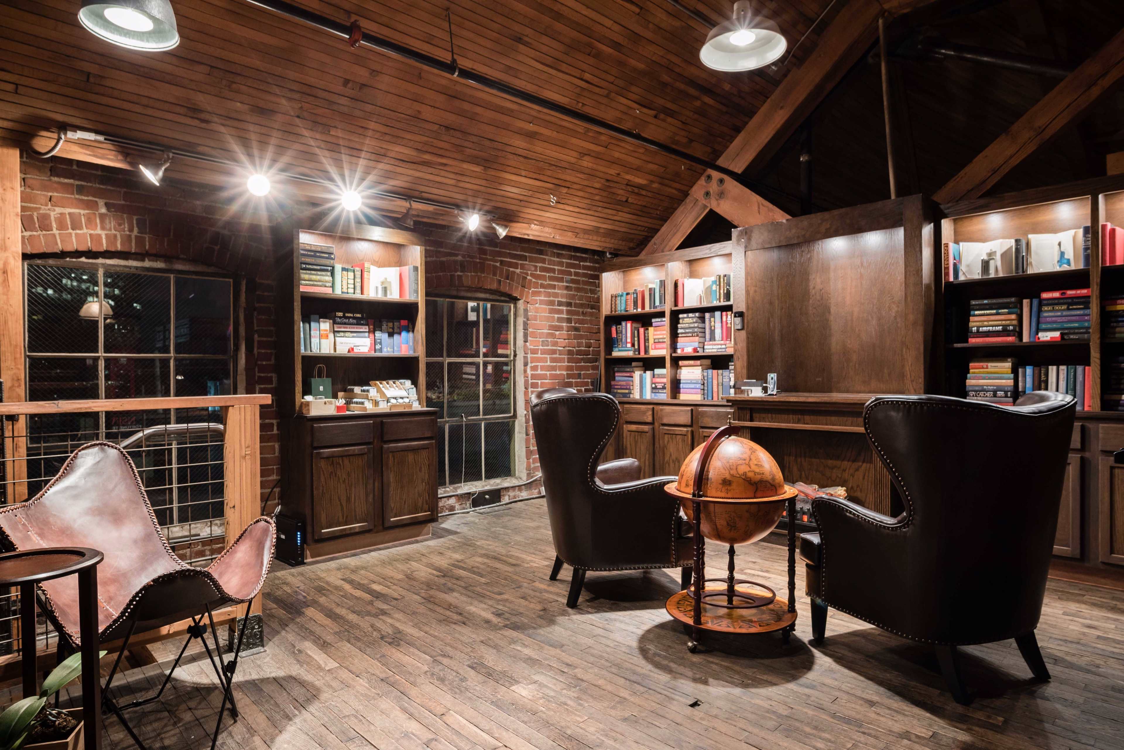 The image shows a cozy library with wooden shelves filled with books, two leather chairs, and a globe on a wooden stand, all set in a space featuring brick walls and a wooden ceiling.