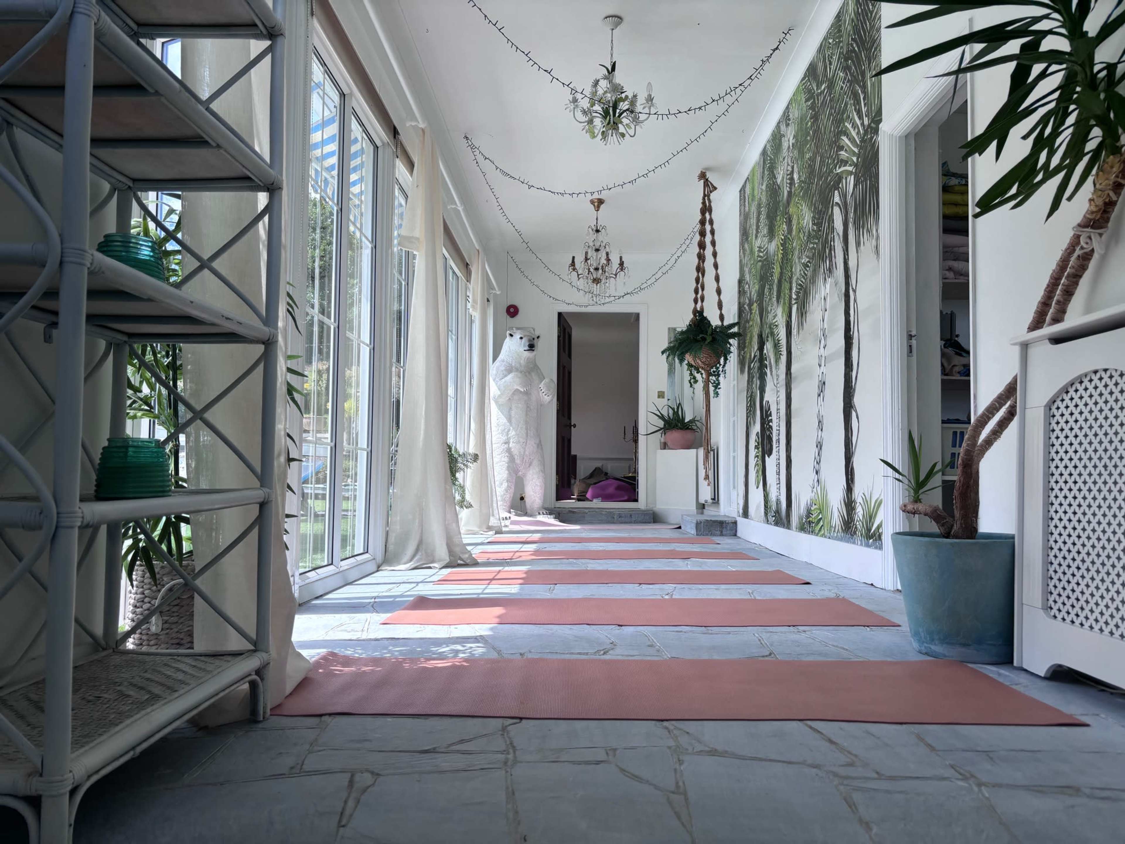 A long hallway features yoga mats on a tiled floor, flanked by large windows, plants, and a mural of palm trees.