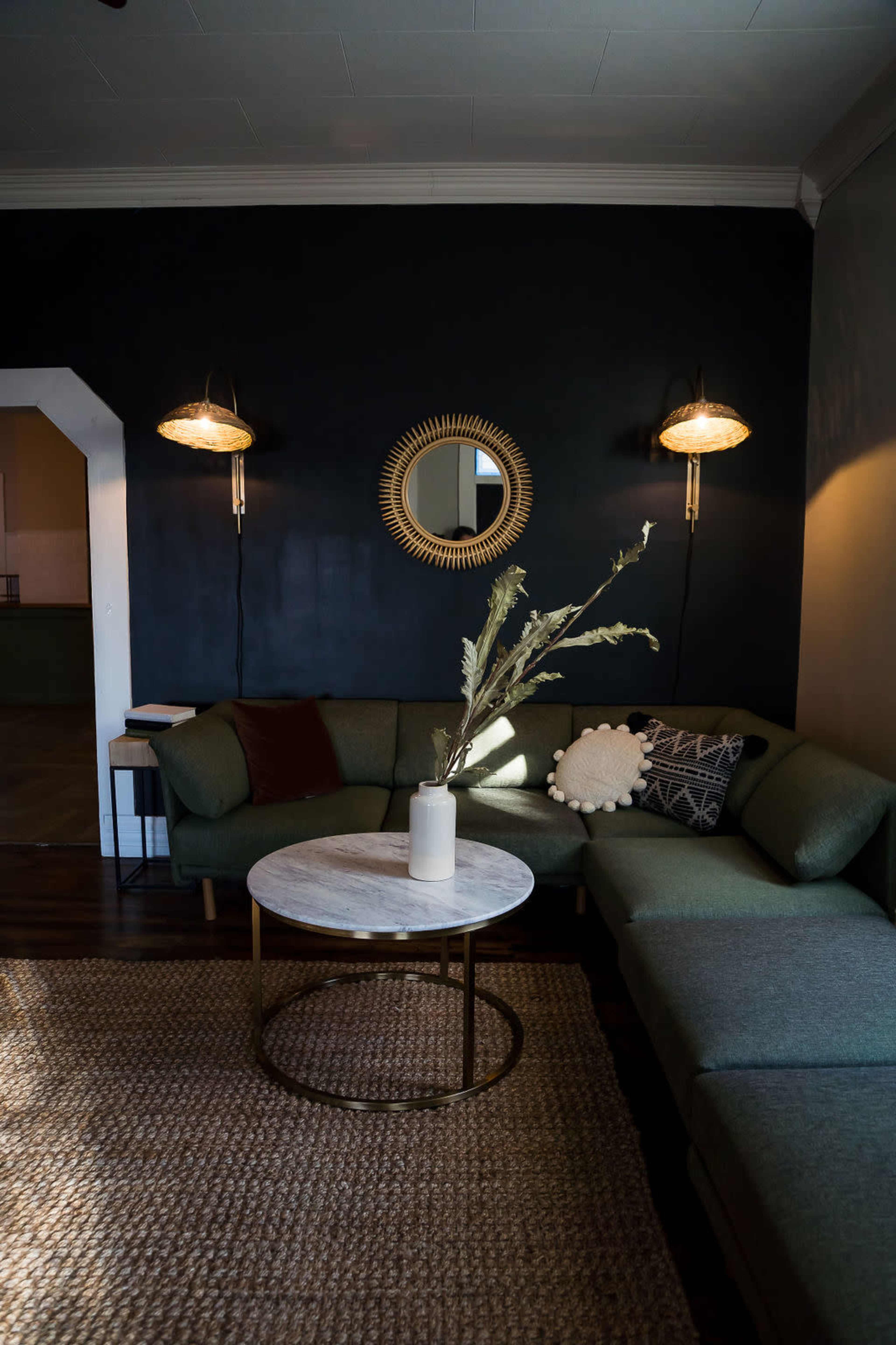 A cozy living room featuring a green sectional sofa, a round marble coffee table with a vase, and wall sconces illuminating a dark wall.