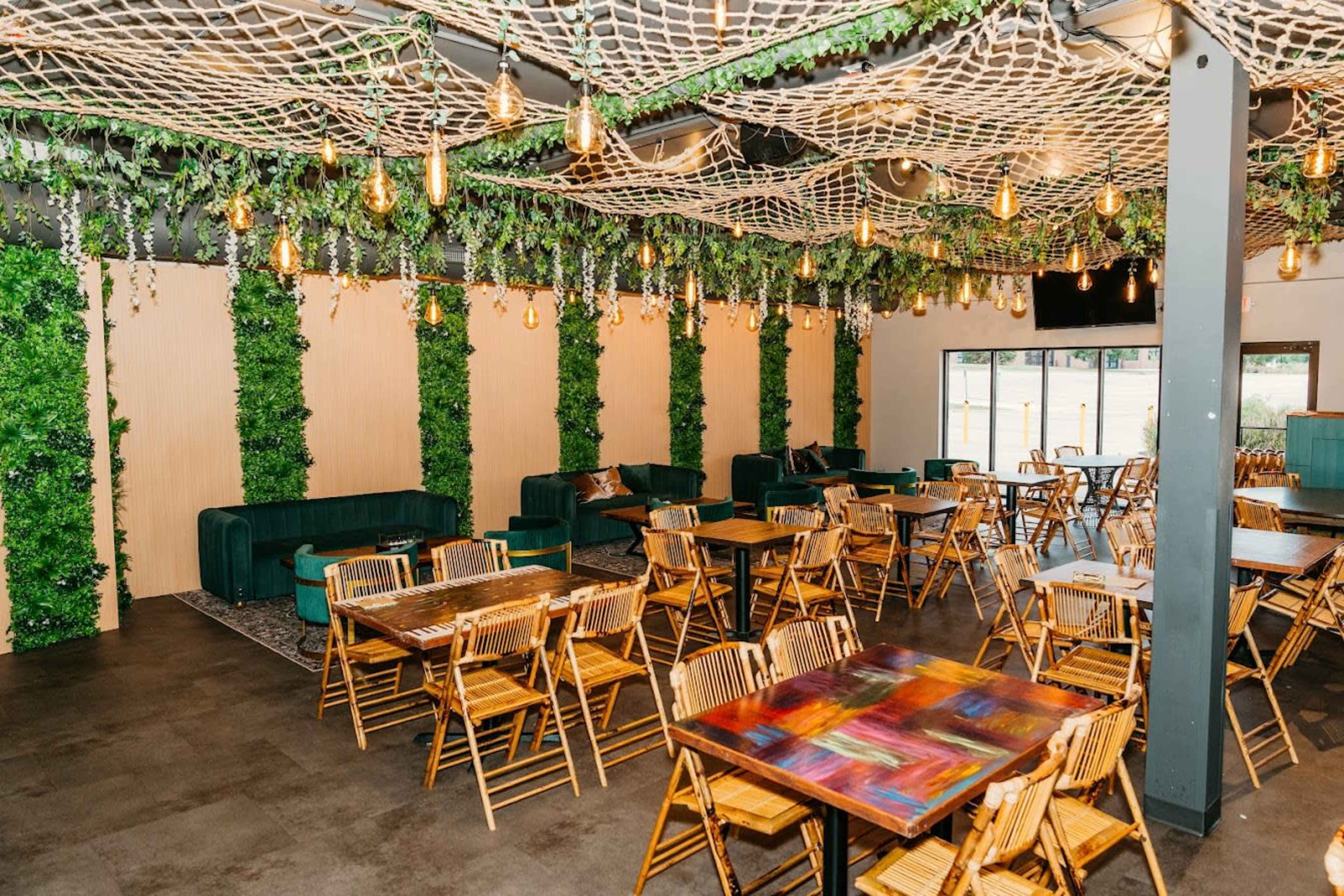 The image shows a spacious restaurant interior with wooden tables and chairs, surrounded by greenery decor and illuminated by hanging lights.