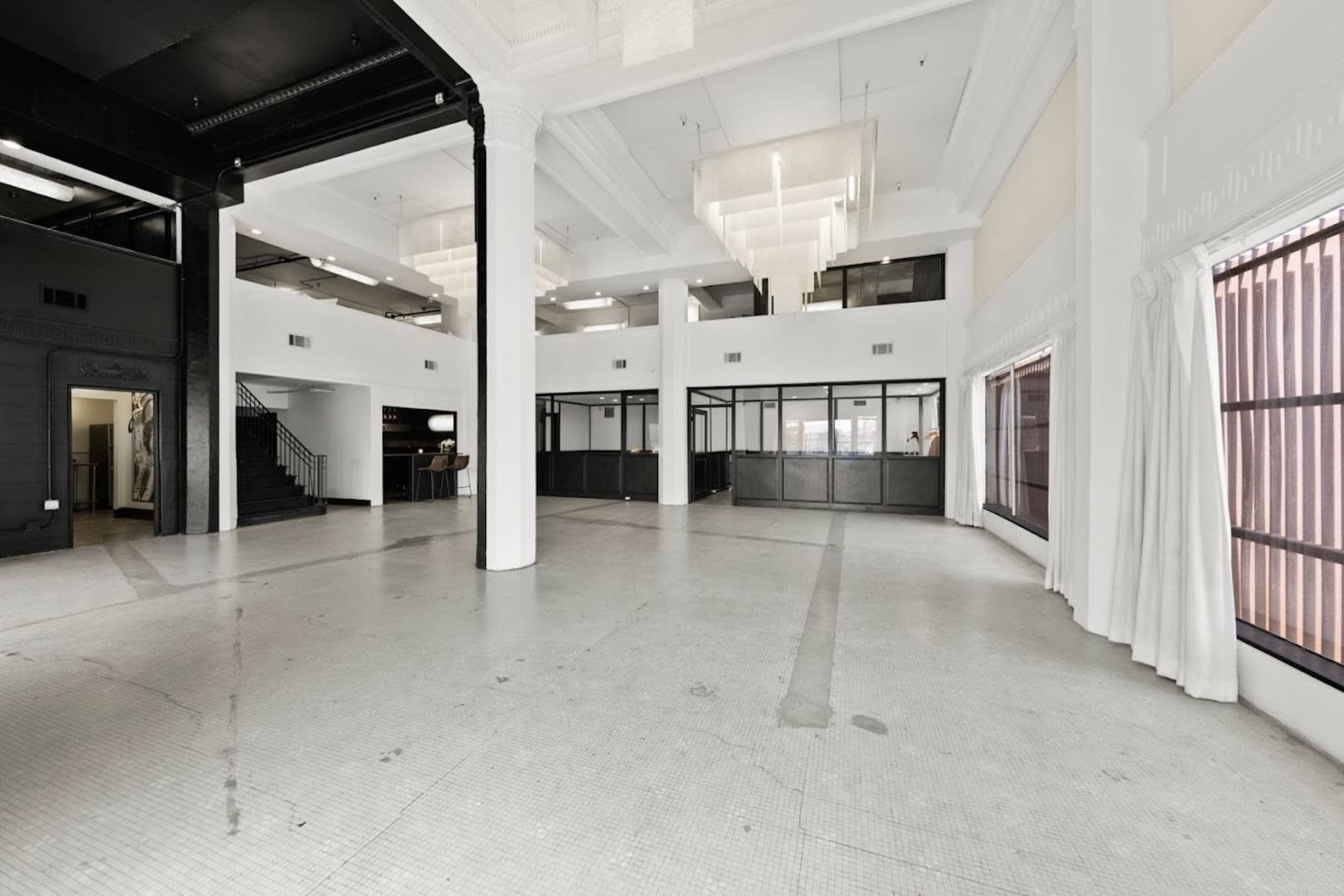 The image shows a spacious, empty interior of a commercial building with high ceilings, large windows, and a black staircase in the background.