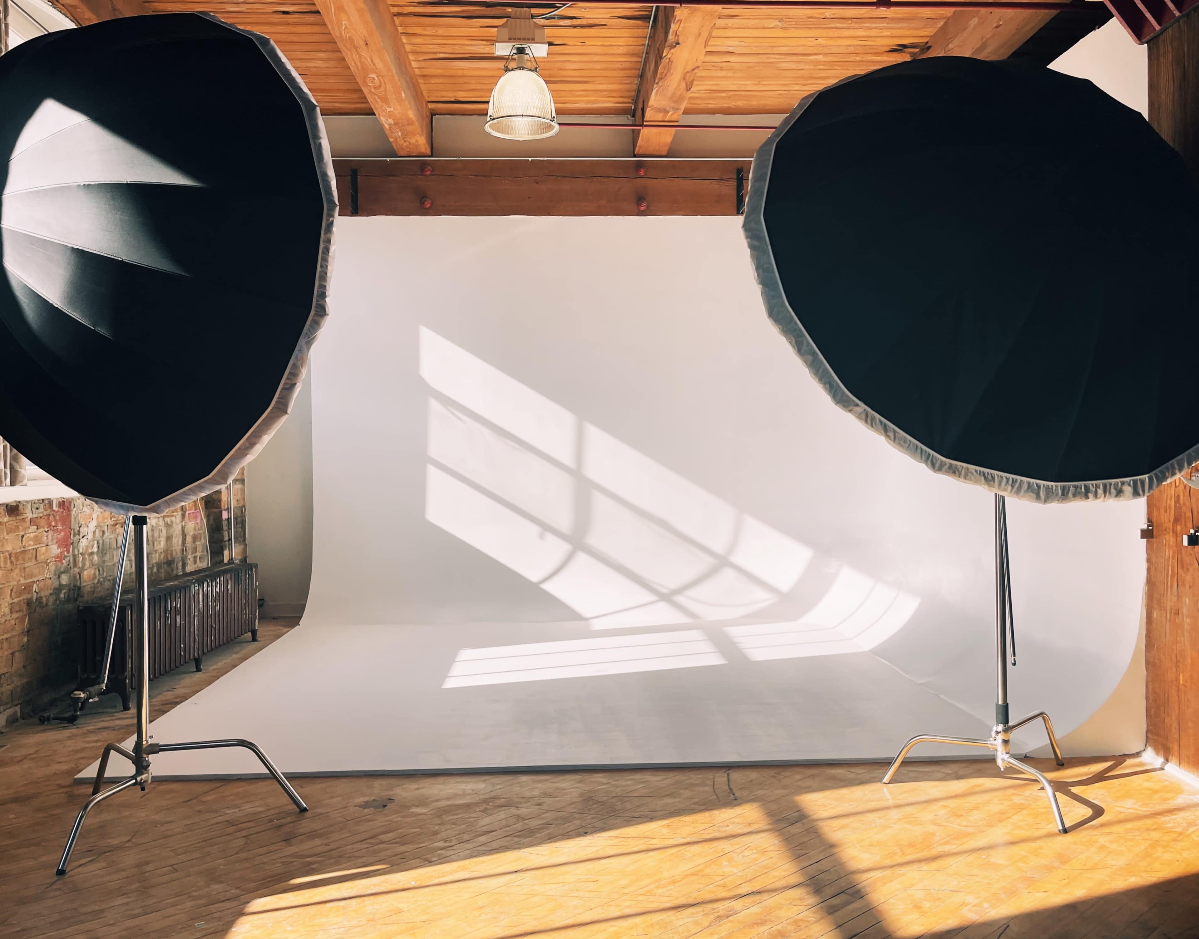 A photography studio with two large softbox lights positioned on either side of a white backdrop, illuminated by natural light streaming through a window.