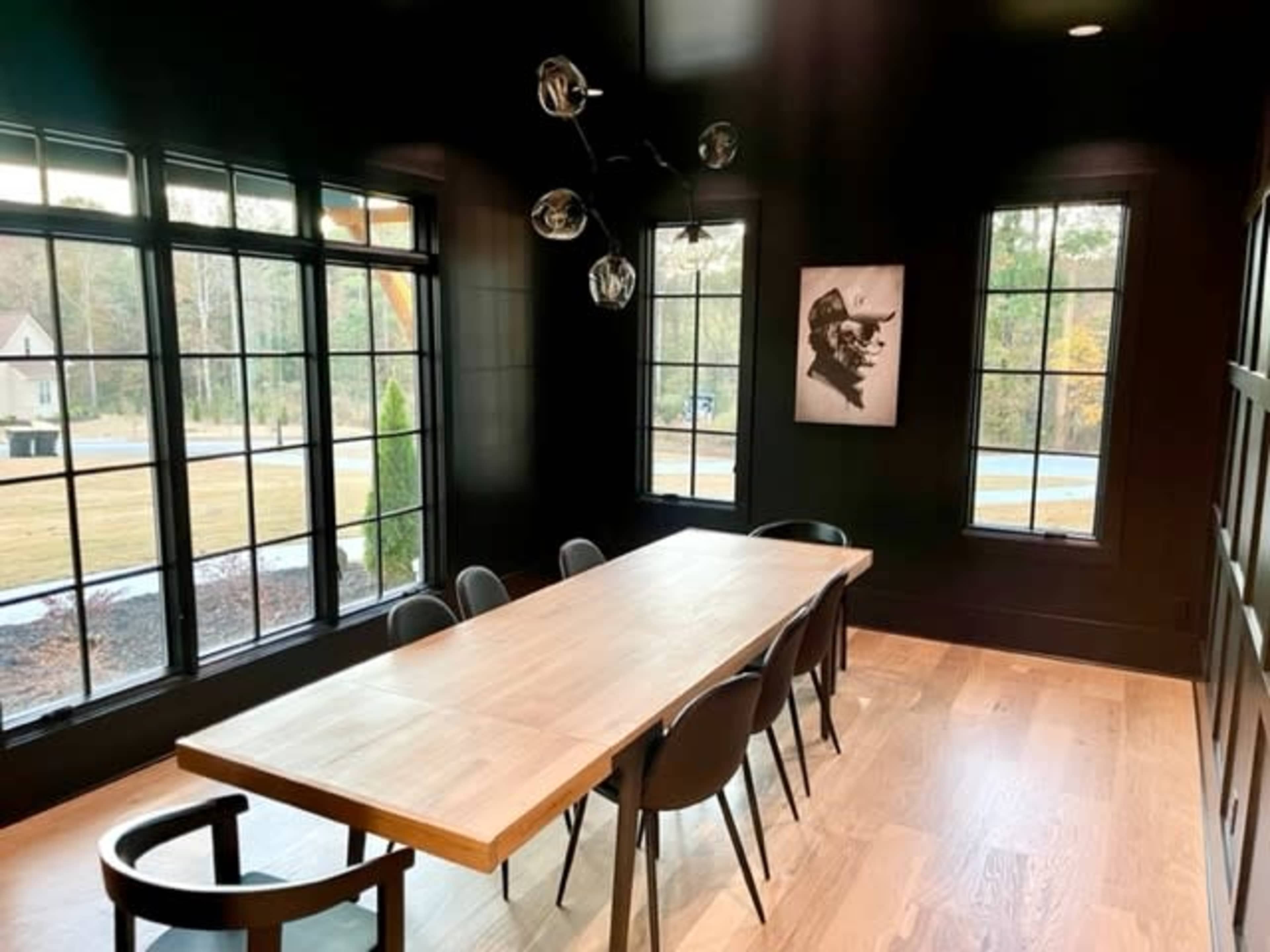 A dining room with a large wooden table and black chairs, illuminated by a modern light fixture, with large windows providing natural light and a view of the landscape outside.