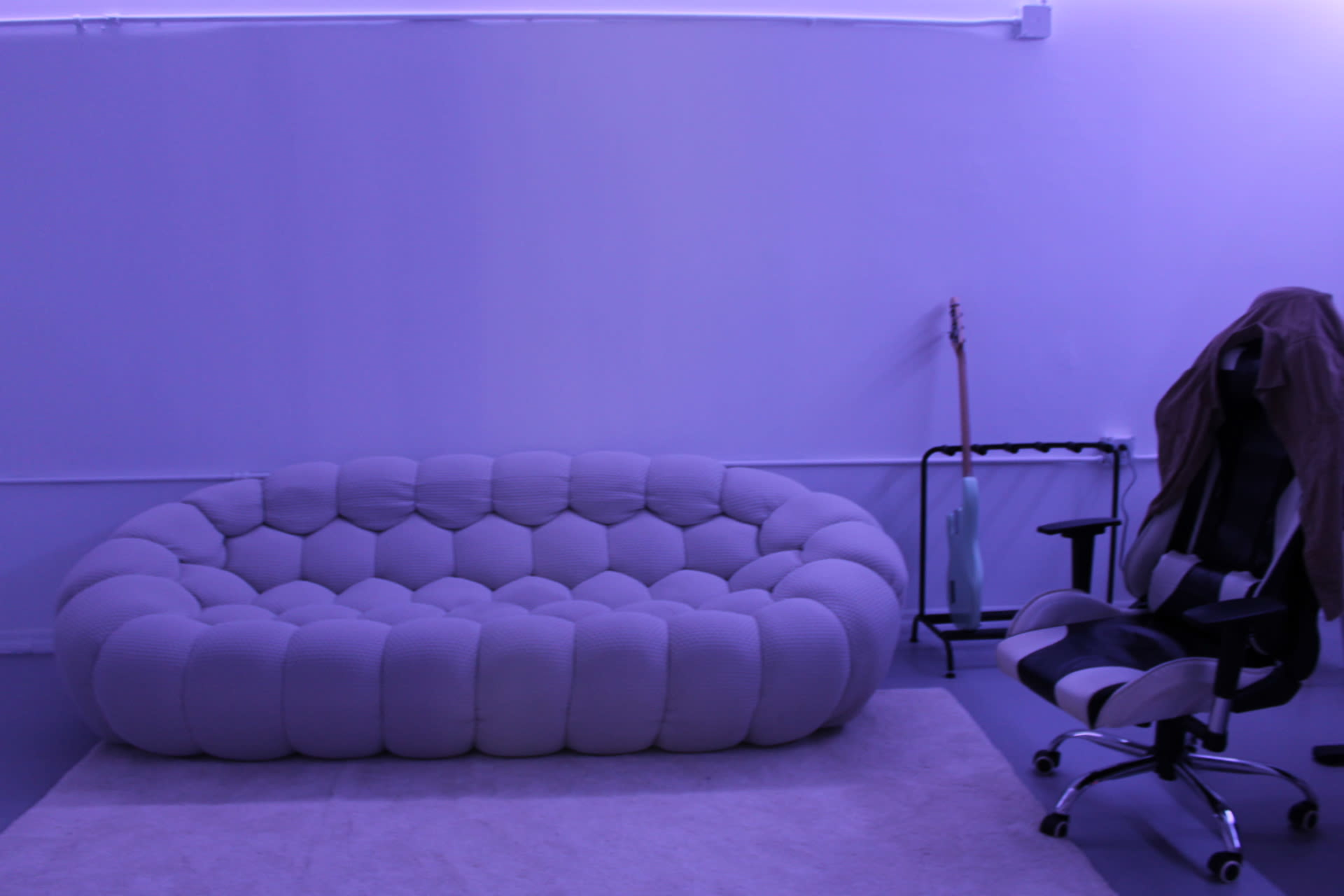 A round, cushioned sofa in a purple-lit room is accompanied by a black and white ergonomic chair and a guitar stand.