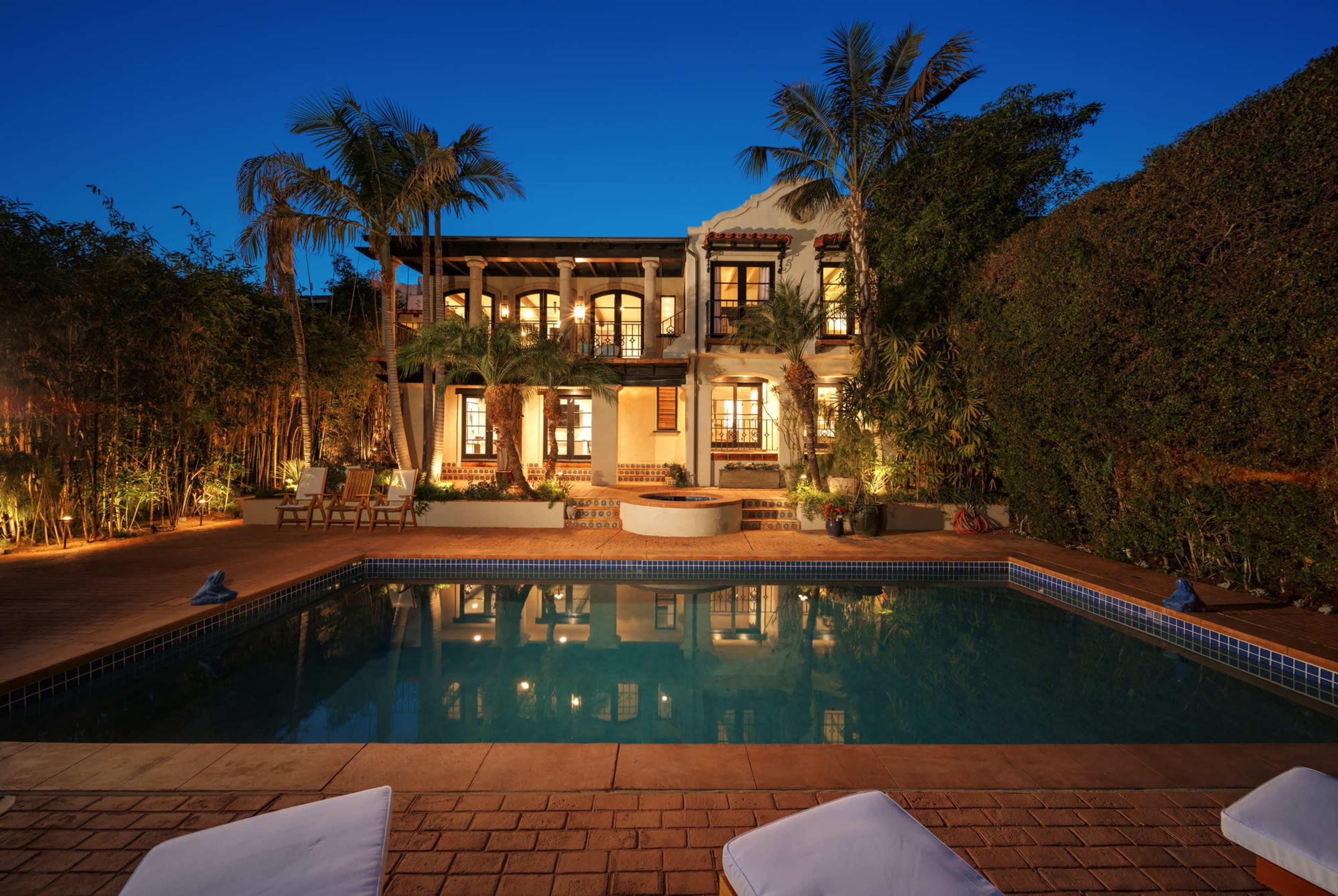 A two-story house with multiple illuminated windows is situated next to a swimming pool surrounded by lush greenery and palm trees.