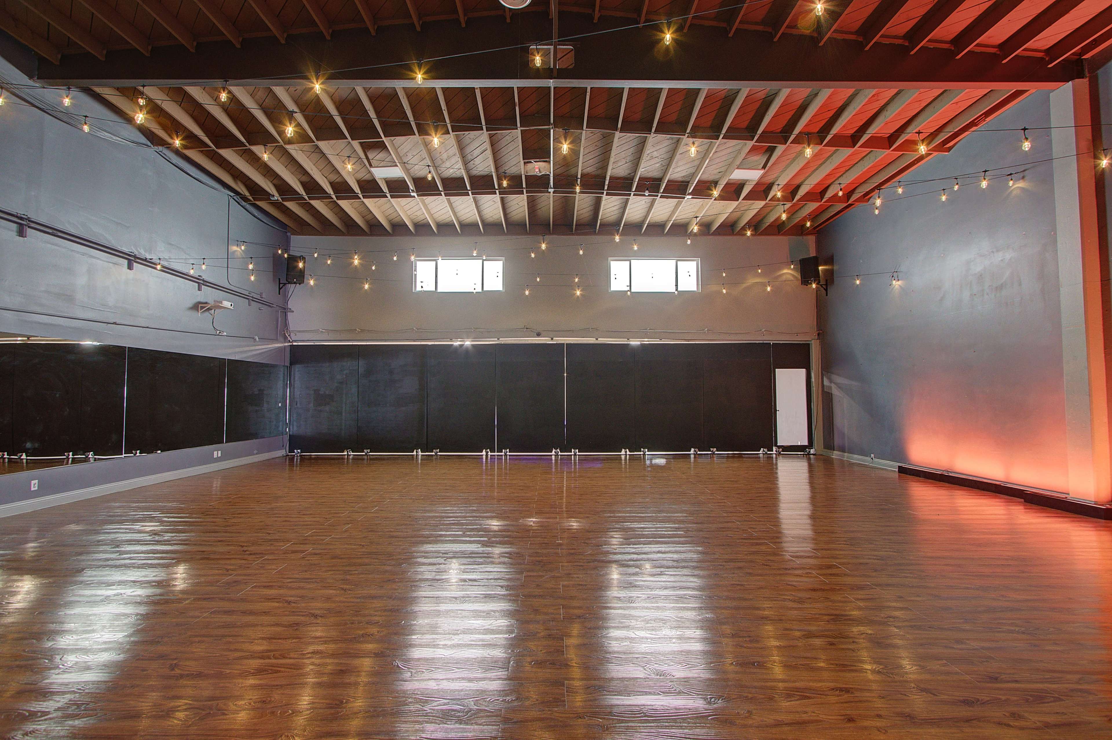 A spacious dance studio with polished wooden floors, mirrors along one wall, and string lights hanging from the ceiling.