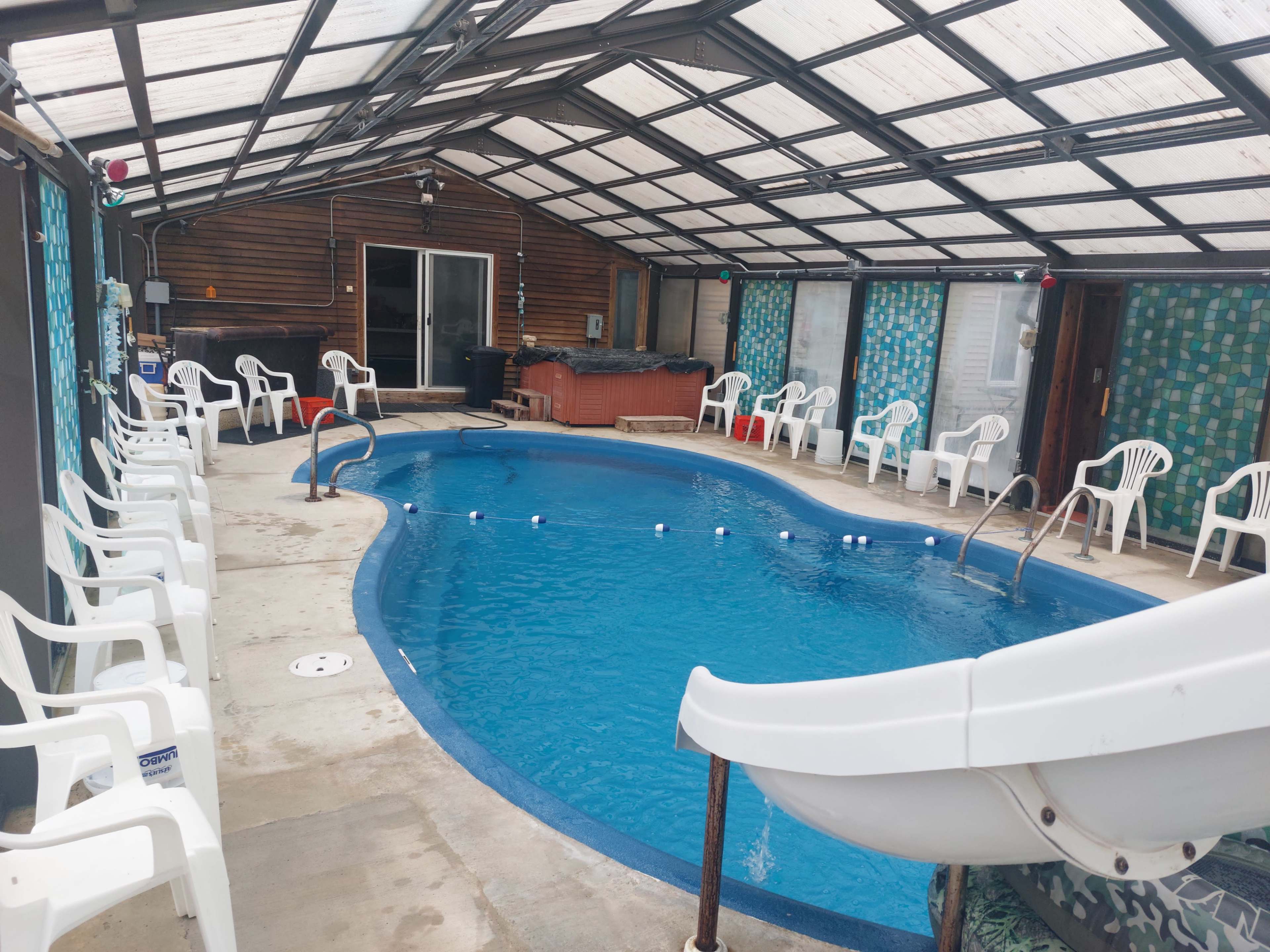 An indoor swimming pool area with a blue pool, a white waterslide, several white plastic chairs arranged around the perimeter, and a hot tub in the corner.