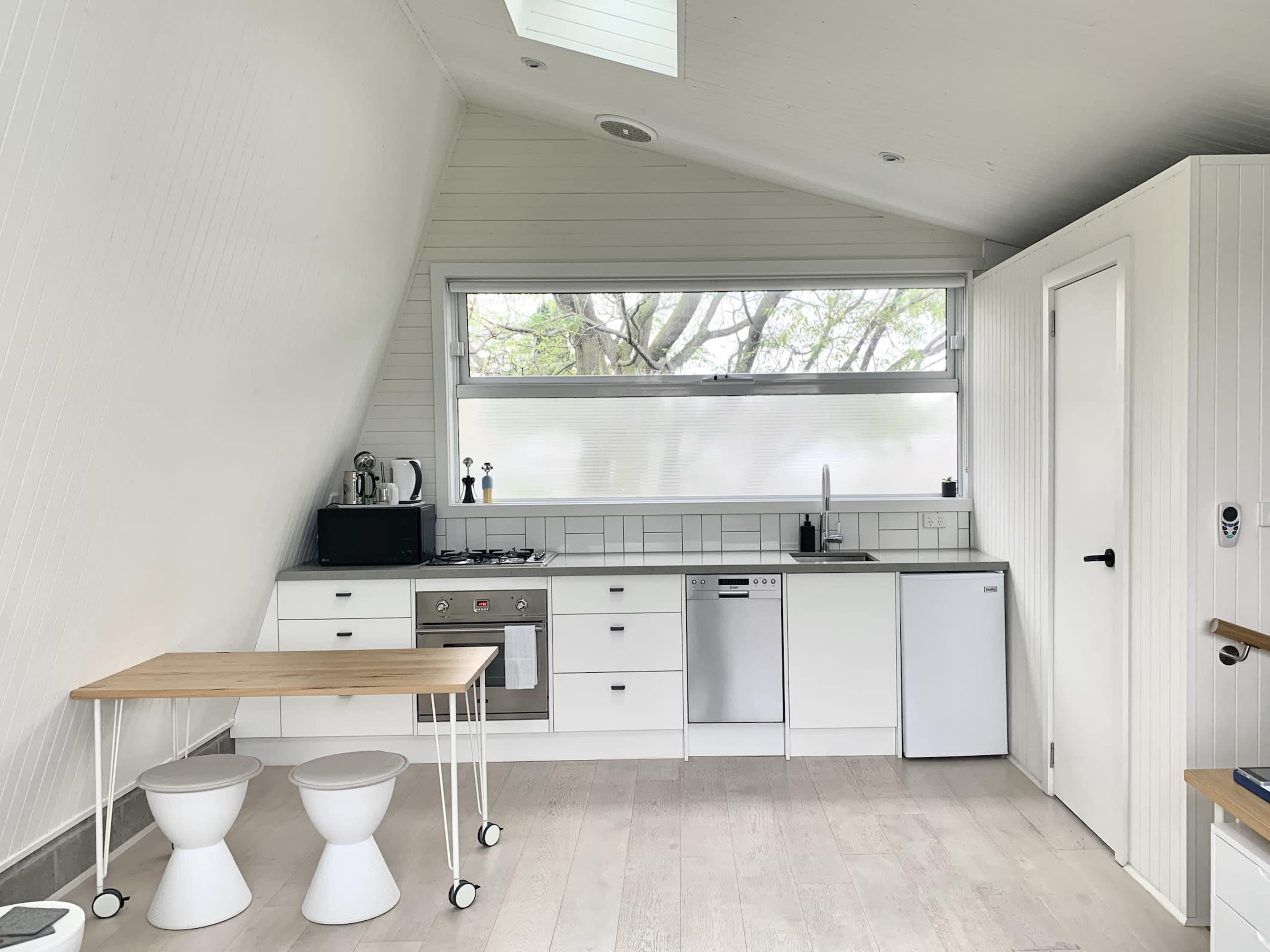 A modern, minimalist kitchen with white cabinetry, stainless steel appliances, and a small wooden dining table with two white stools.