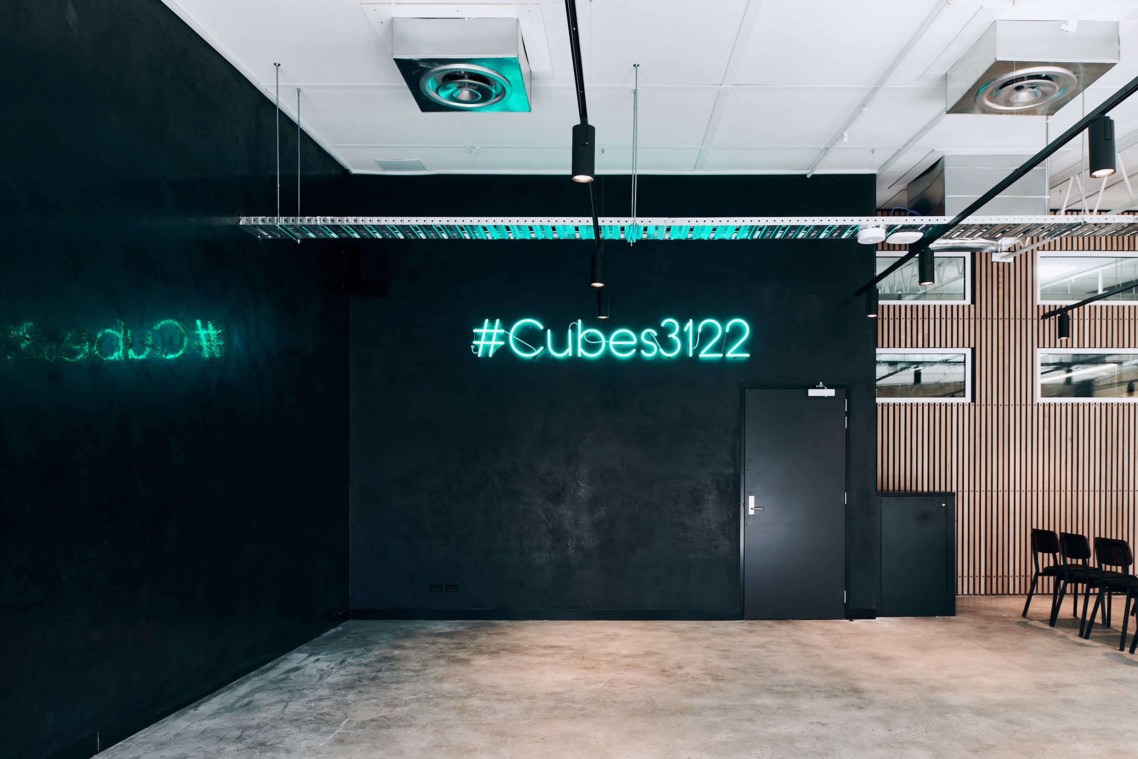 The image shows an interior space with a dark wall featuring a neon sign that reads "#Cubes3122" and a door leading to another area.