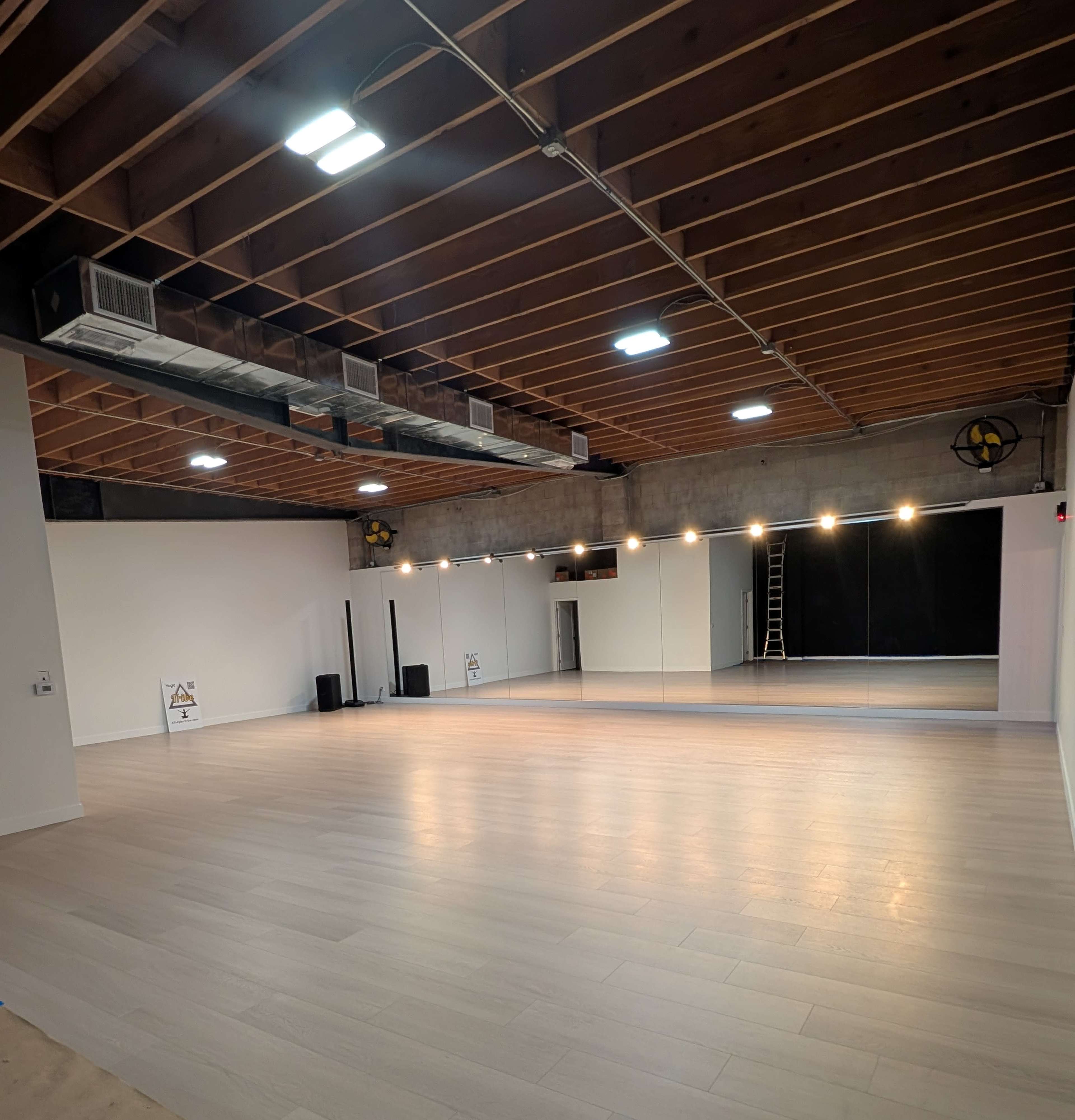 The image shows a spacious, well-lit studio with wooden floors, exposed ceiling beams, and a large mirror on one wall.