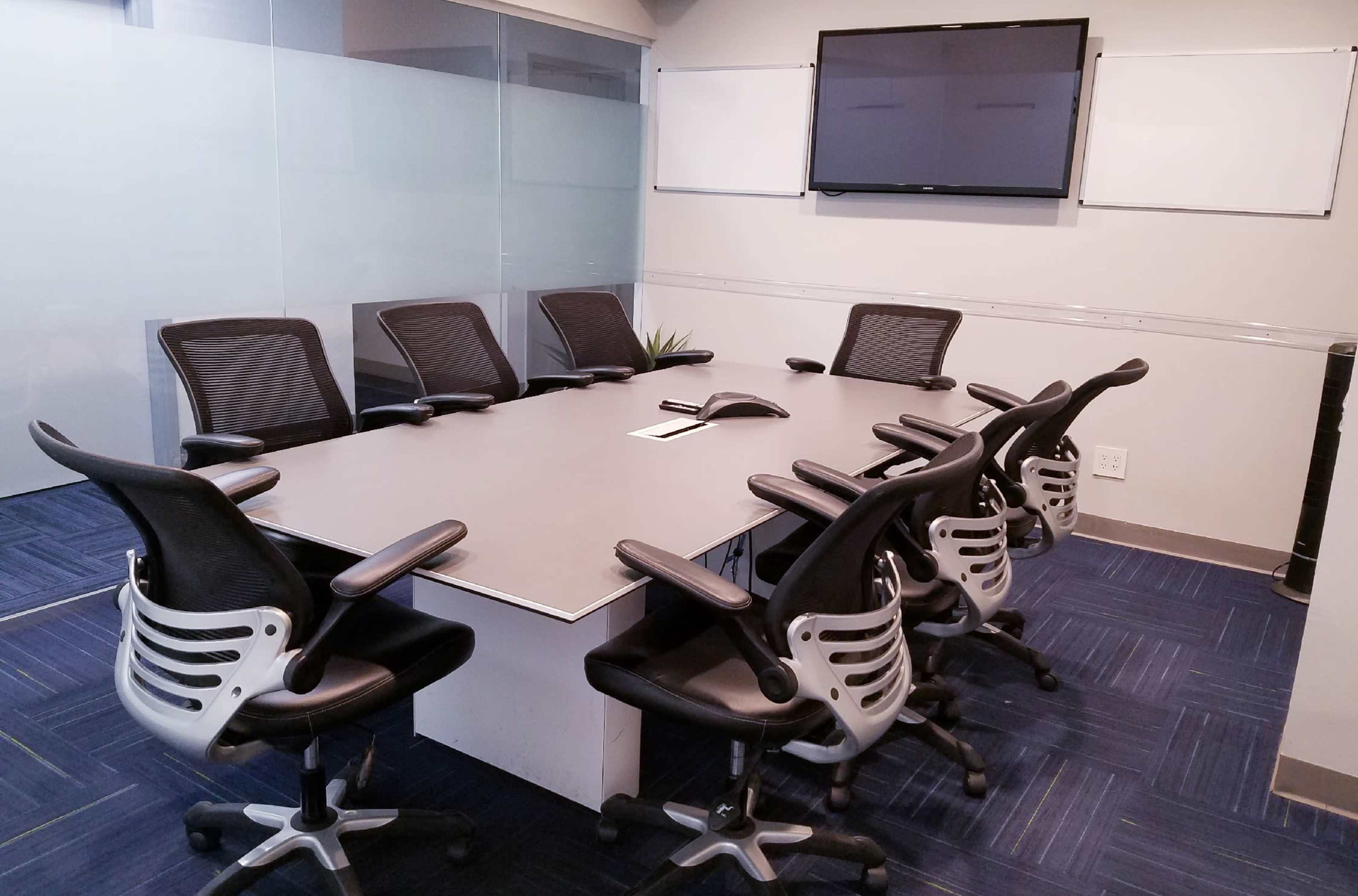 The image shows a modern conference room with a long table, eight ergonomic chairs, a television mounted on the wall, and whiteboards.