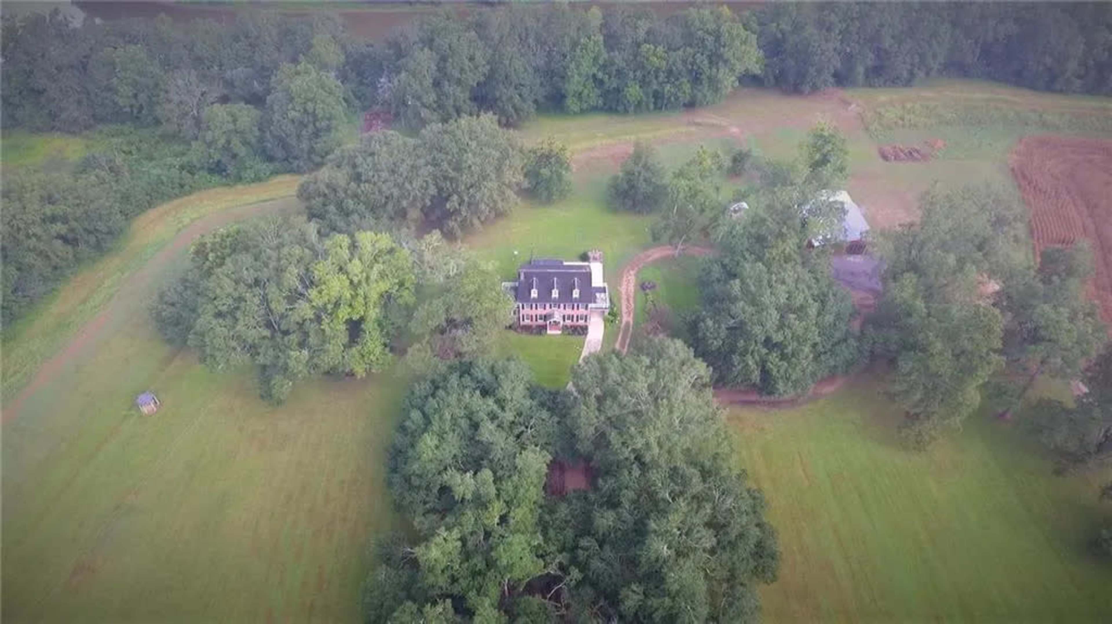 The image shows a large house surrounded by trees and fields, situated in a rural area.