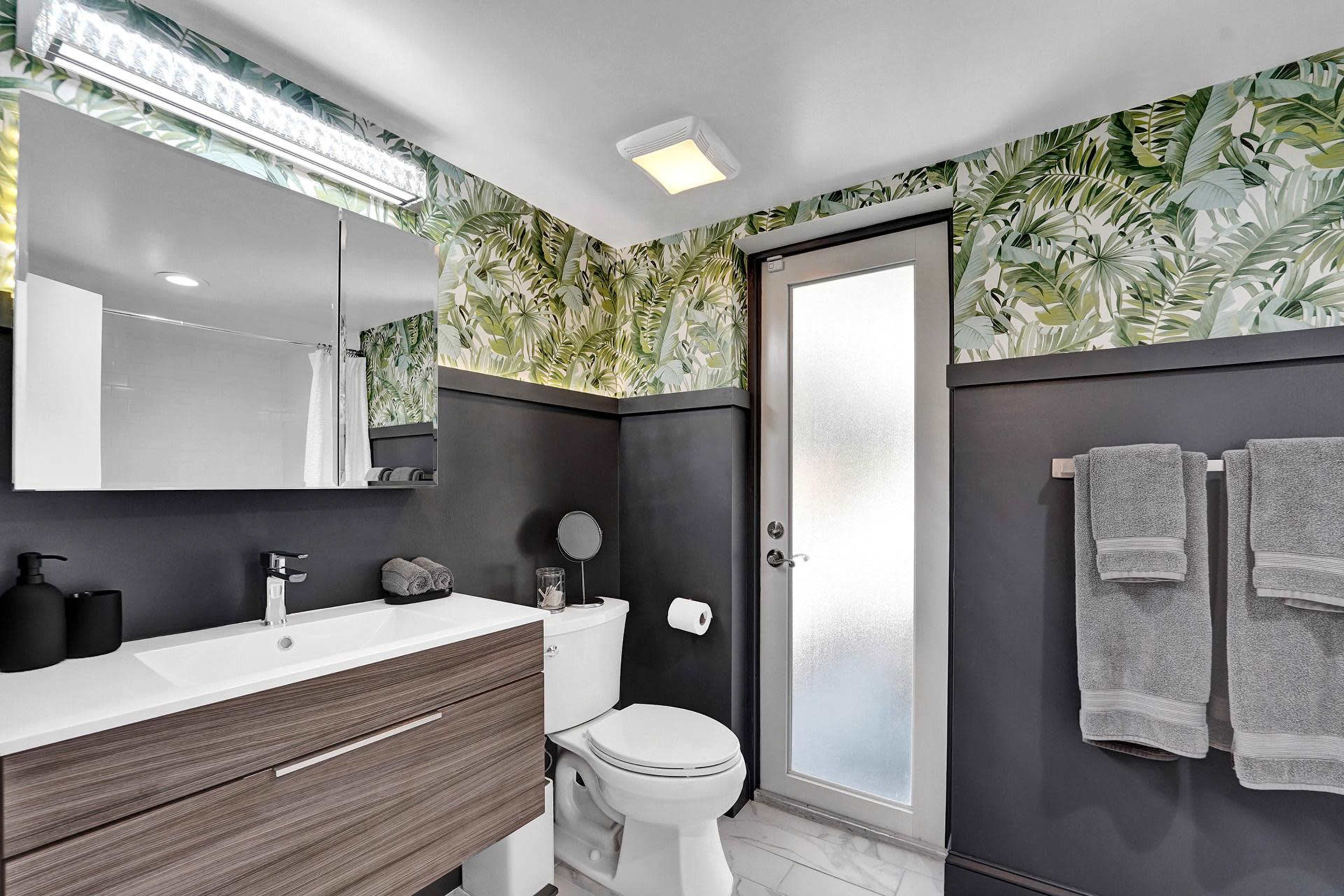 A modern bathroom features a wooden vanity, a frosted glass door, and tropical-themed wallpaper.