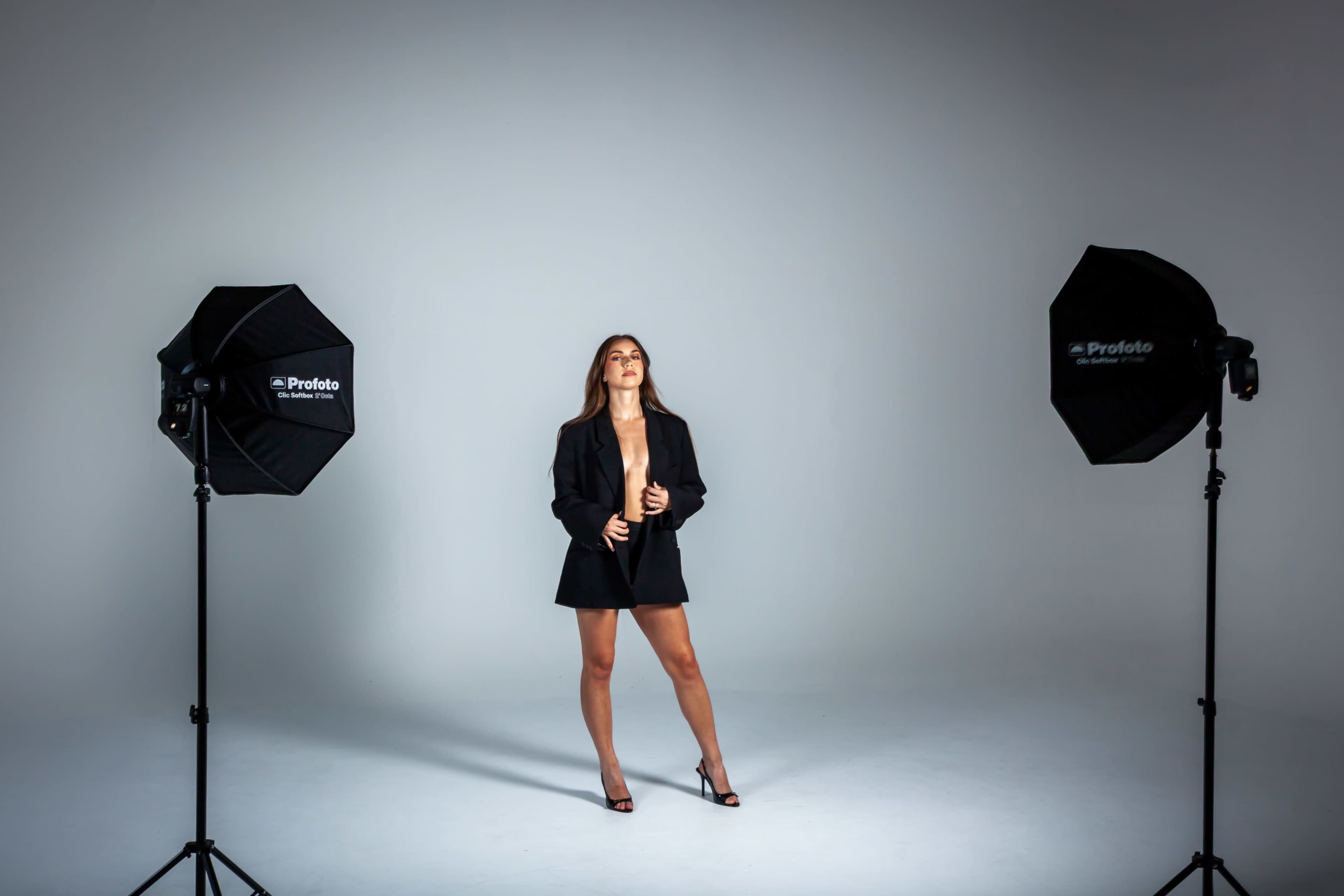 A model stands in a studio setting between two softbox lights, wearing a blazer and high heels.