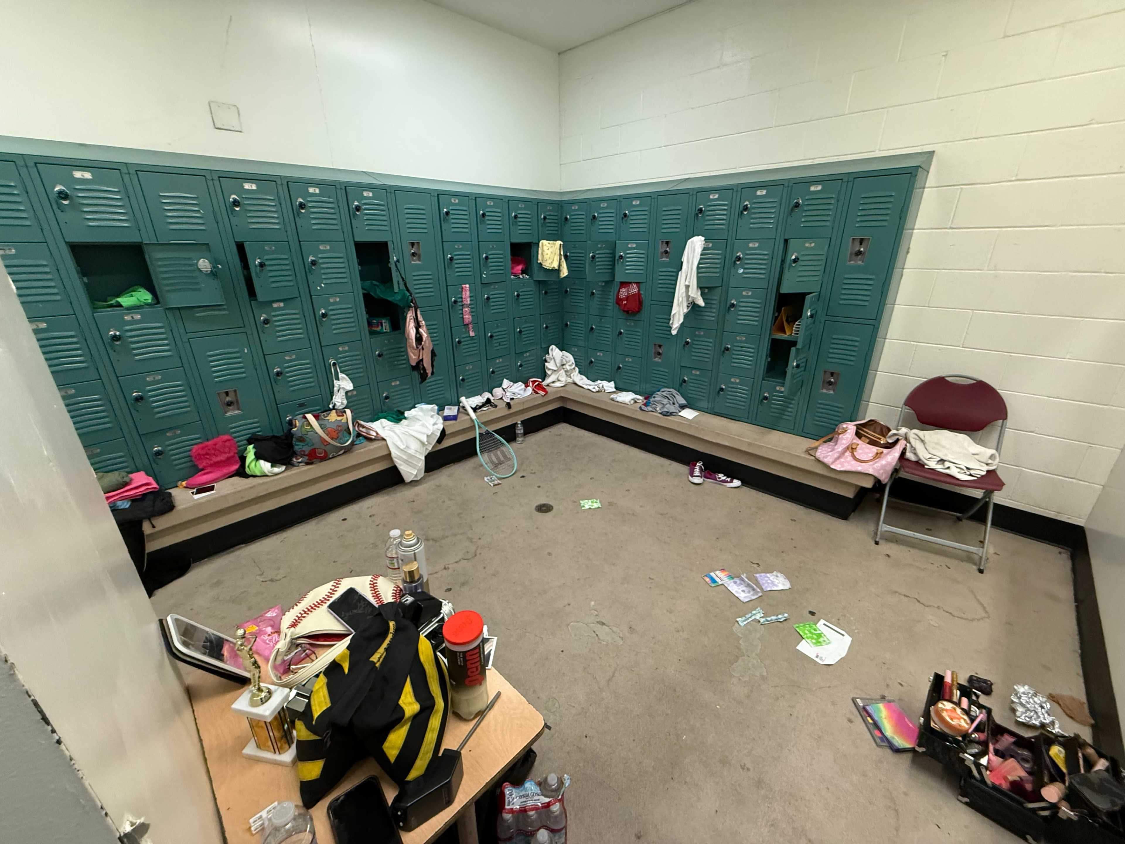 The image shows a cluttered locker room with open lockers, scattered clothing, and personal items across the floor and benches.