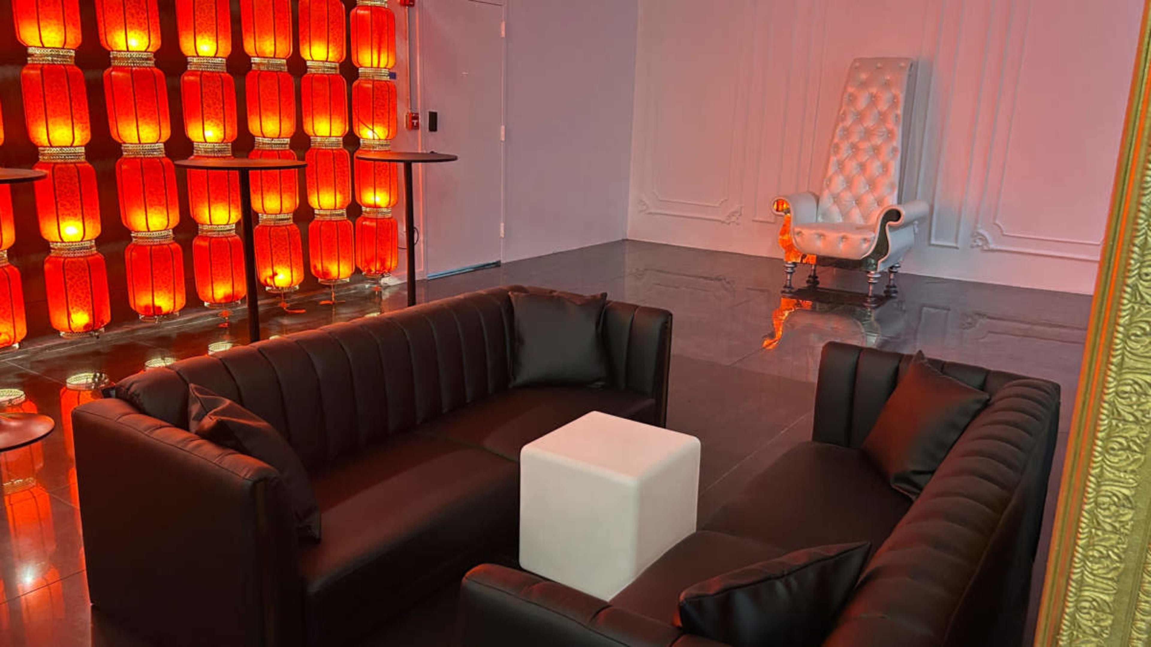 The image shows a modern lounge area featuring black leather sofas, a white cube table, and a large, ornate white chair against a backdrop of illuminated red lanterns.