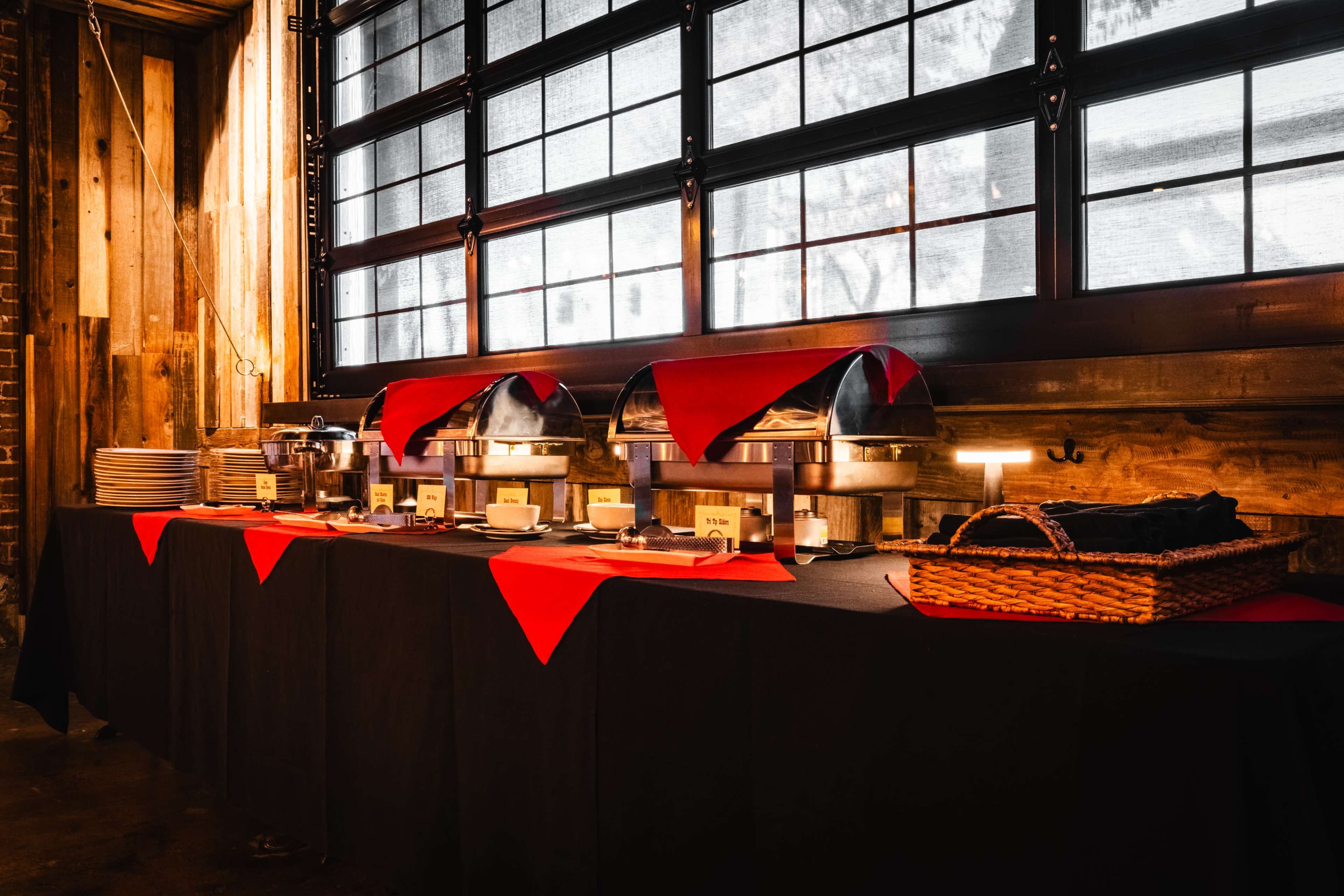 A buffet table with stacked plates and food warmers is set against a backdrop of wooden walls and large windows.