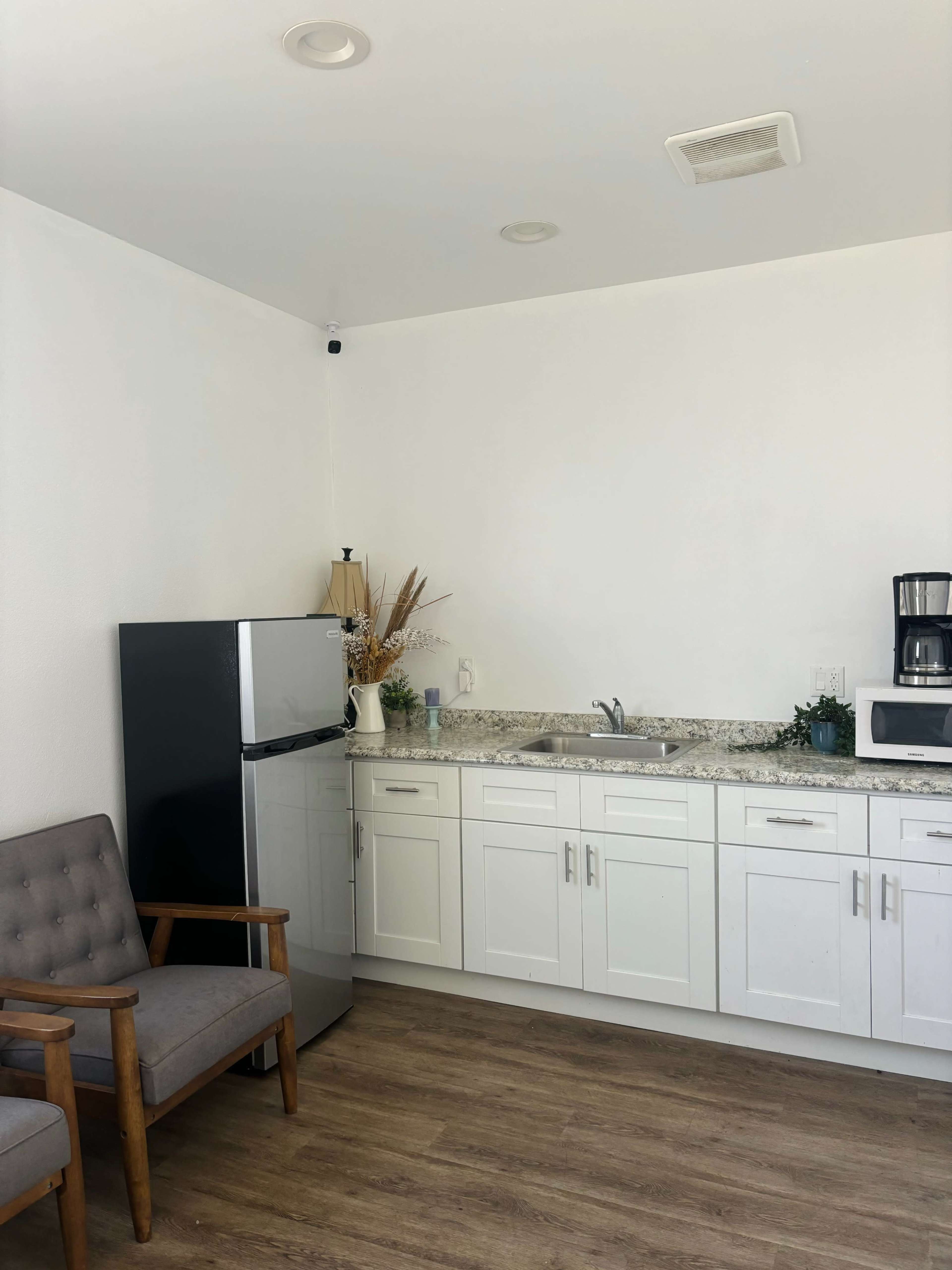 The image shows a modern kitchenette with a black refrigerator, a microwave, and a coffee maker, accompanied by a small gray chair and decor elements on the counter.