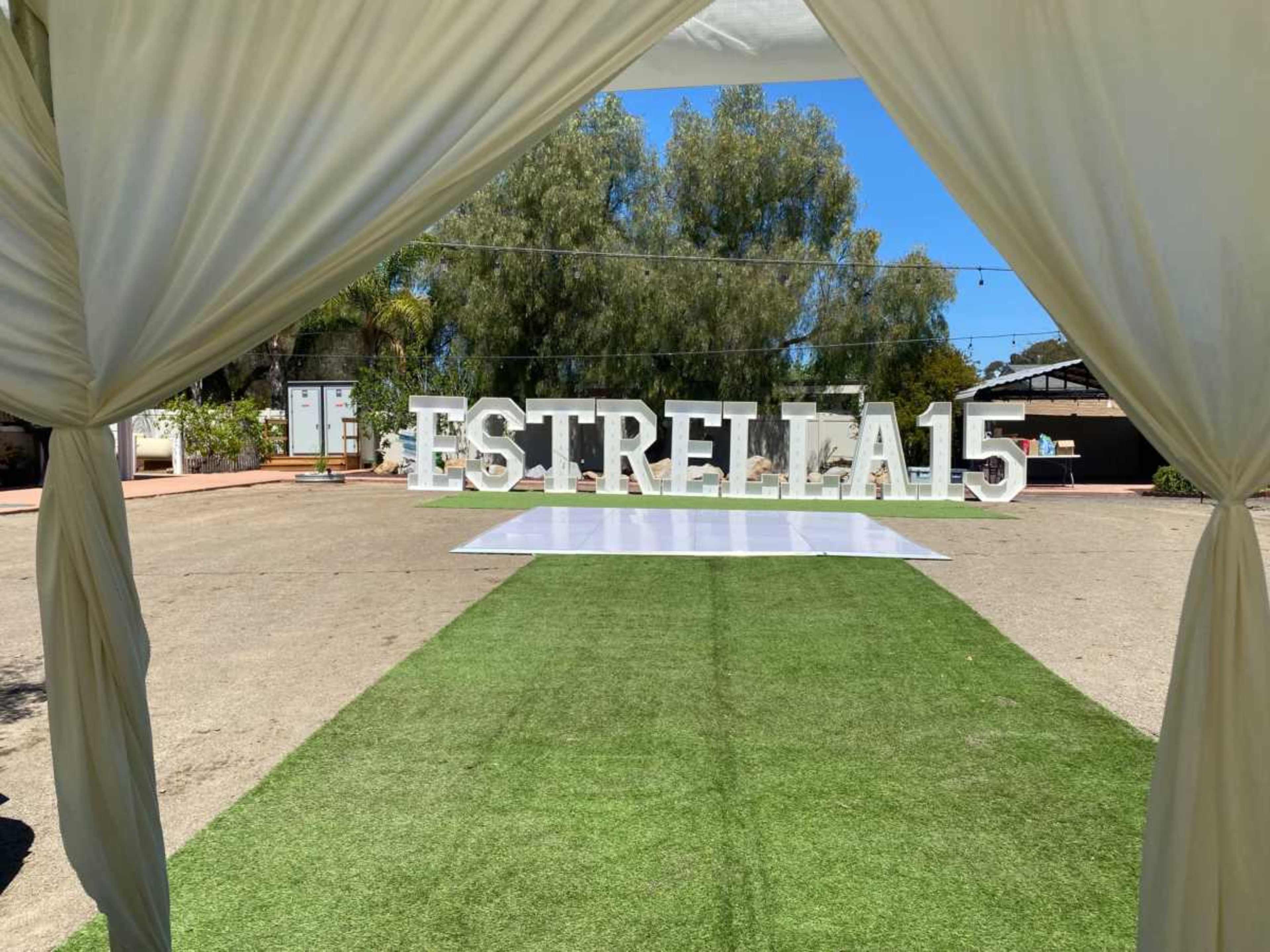 The image shows a party setup featuring large white letters that spell "ESTRELLA 15" on a grassy area, framed by a tent drape.
