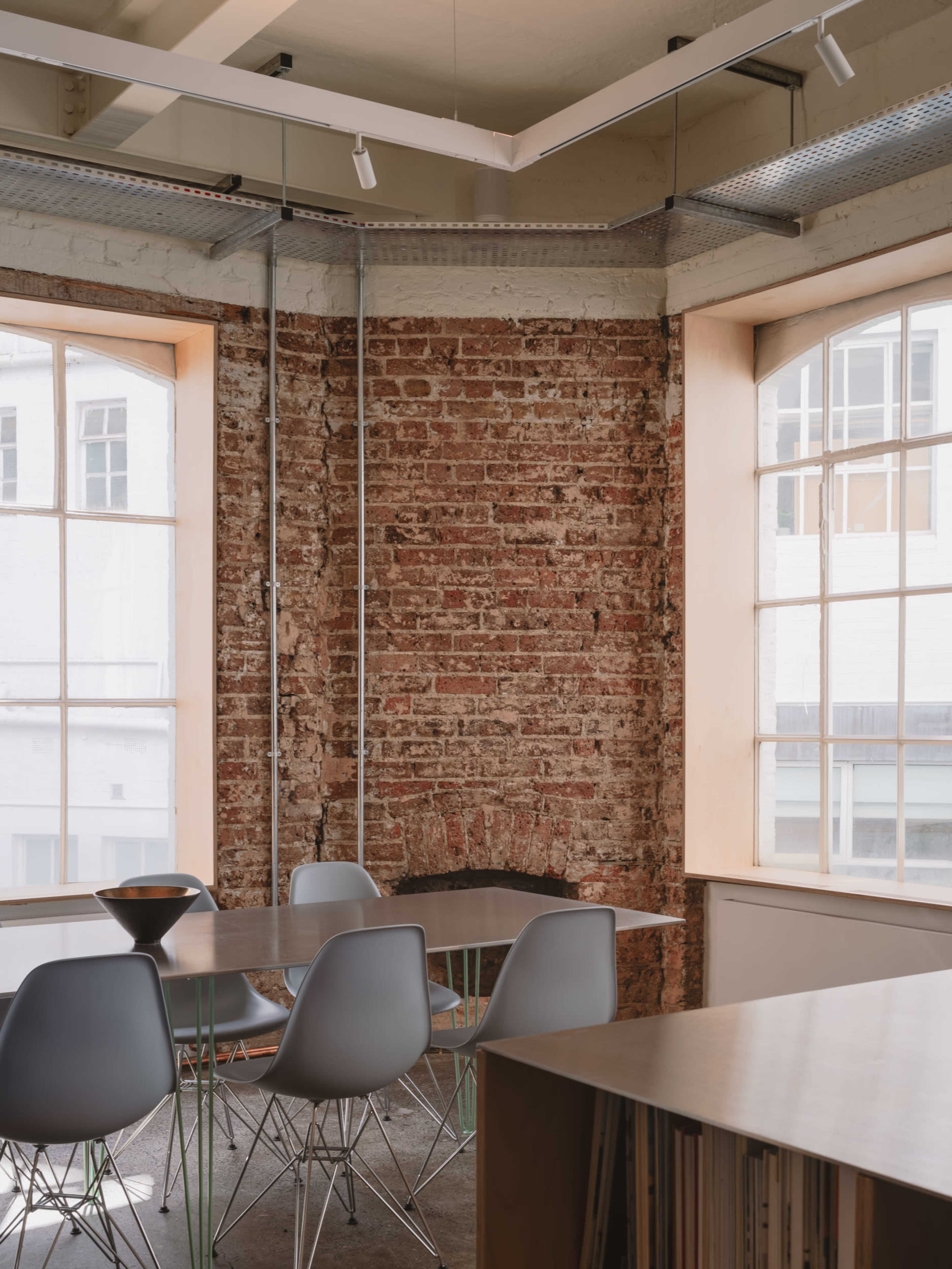 The image shows a modern workspace featuring a table with gray chairs adjacent to large windows and a textured brick wall.