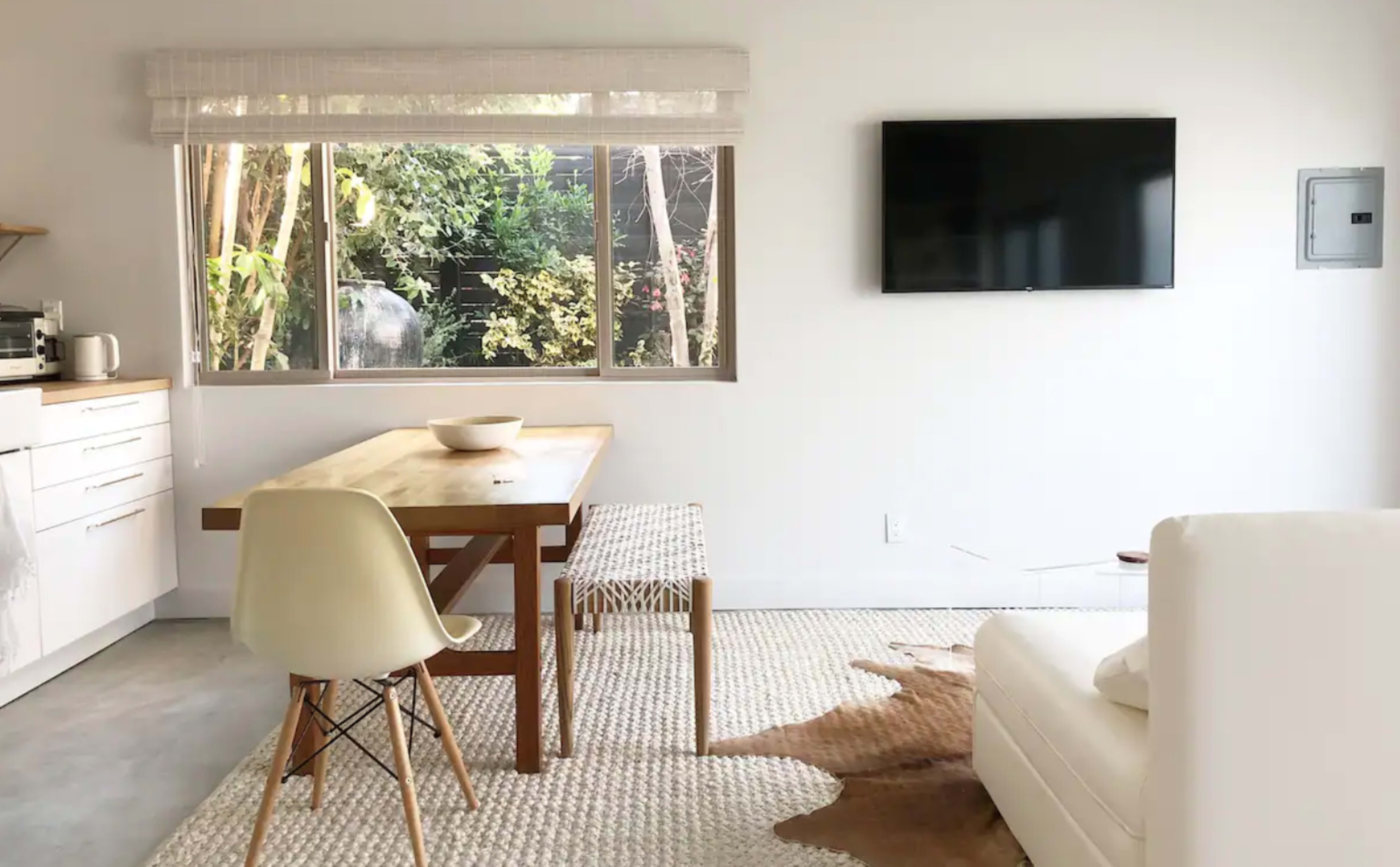 A modern dining area features a wooden table, white chair, and a large window overlooking a lush garden, with a television mounted on the wall.