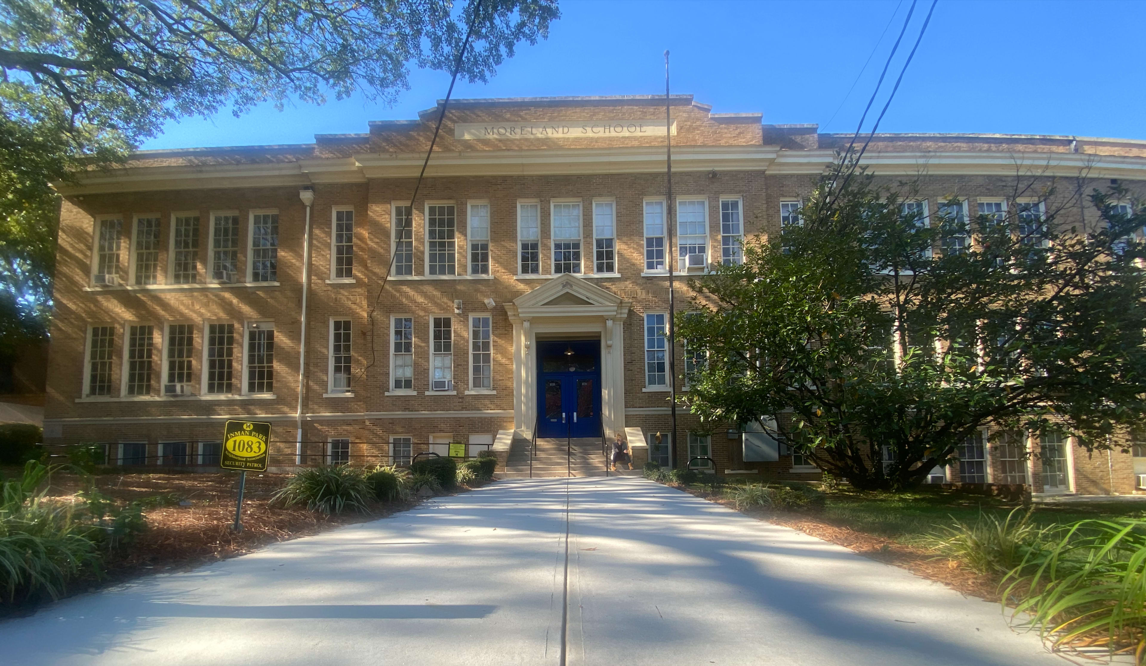 A brick school building with large windows, a central entrance, and a tree-lined pathway leading up to it.