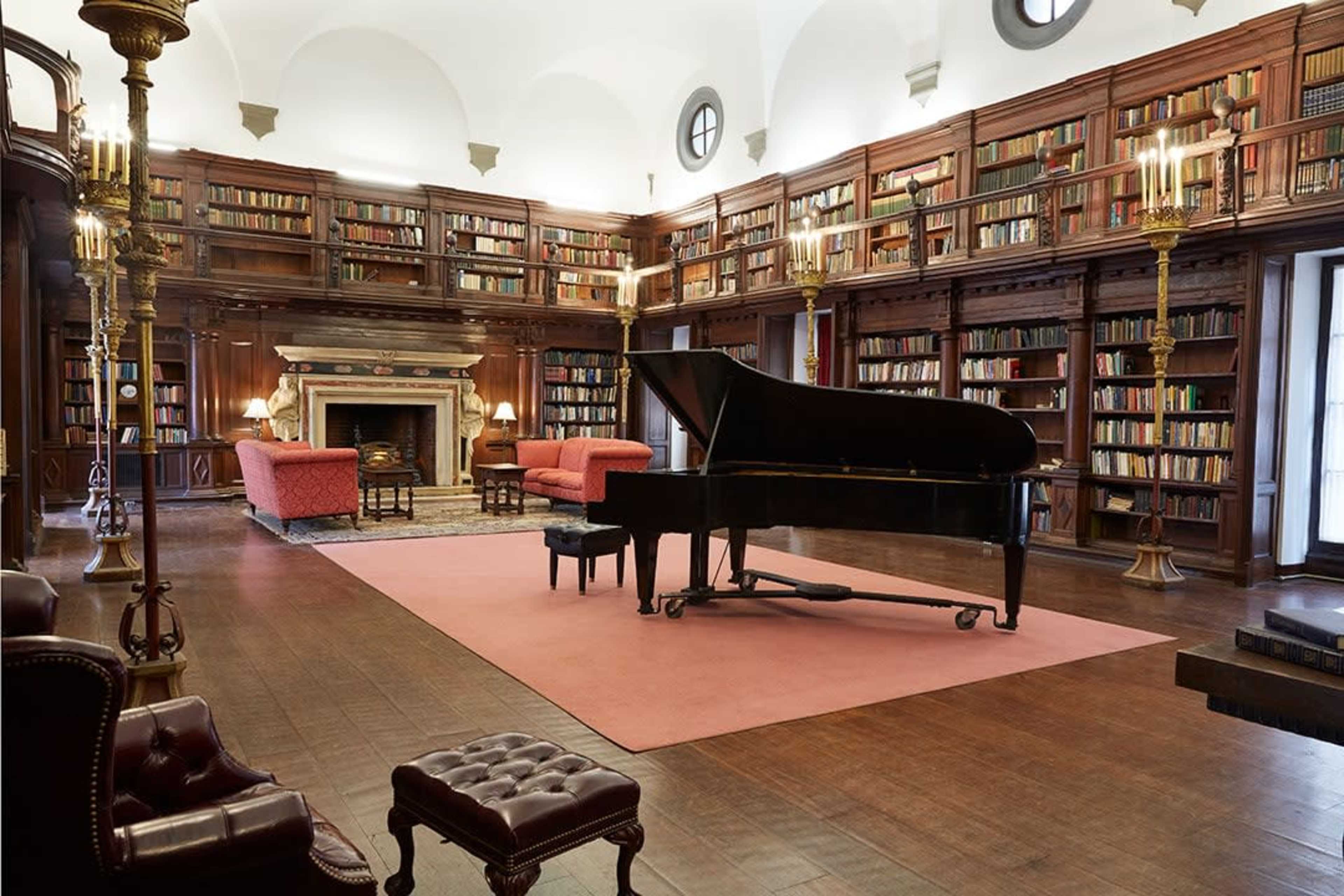 The image shows a spacious library with tall bookshelves filled with books, a grand piano in the center, and elegant seating areas on either side.