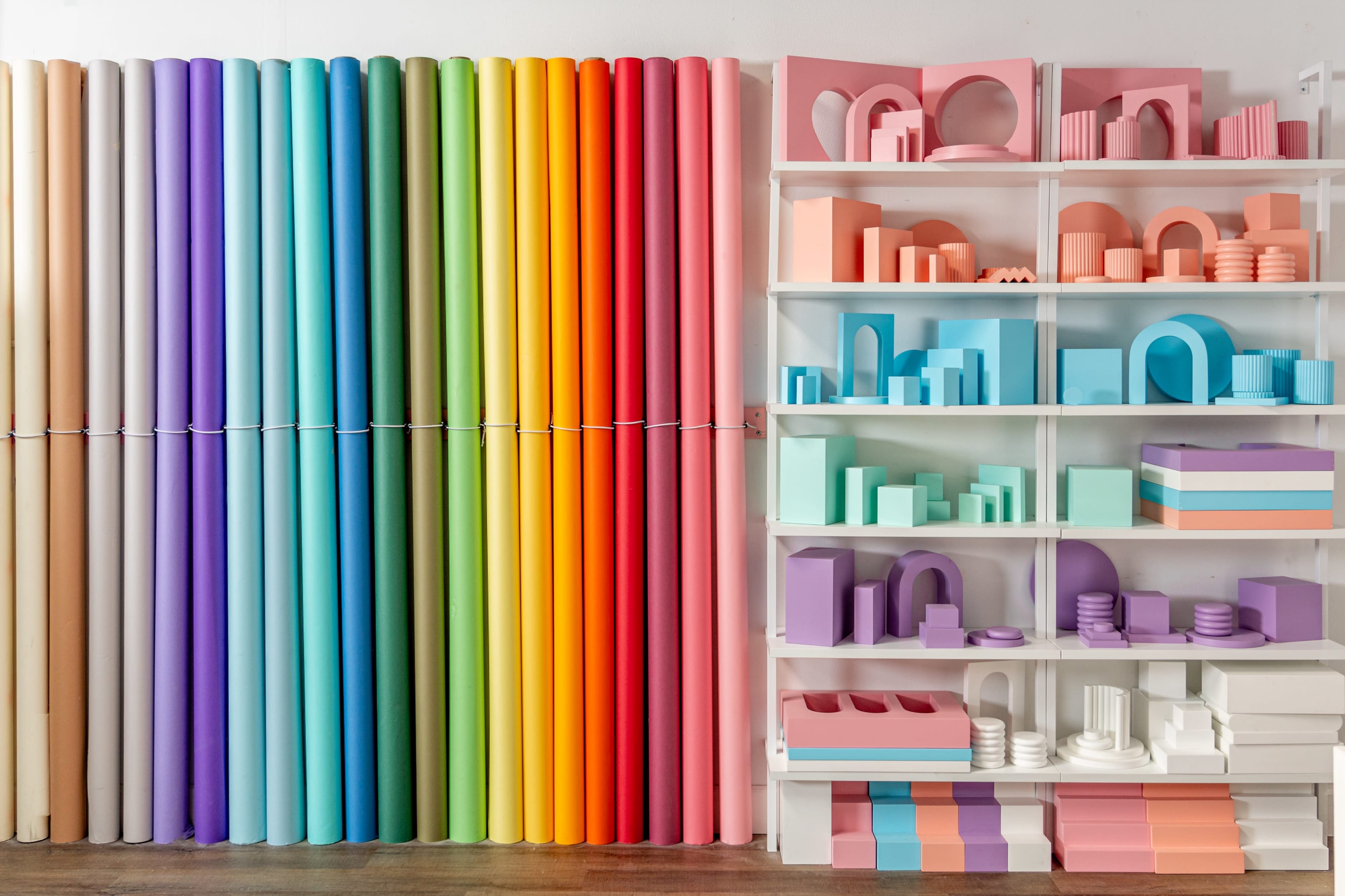 A wall of colorful rolled paper in gradient hues on the left and a white shelf displaying various geometric shapes and objects in pastel colors on the right.