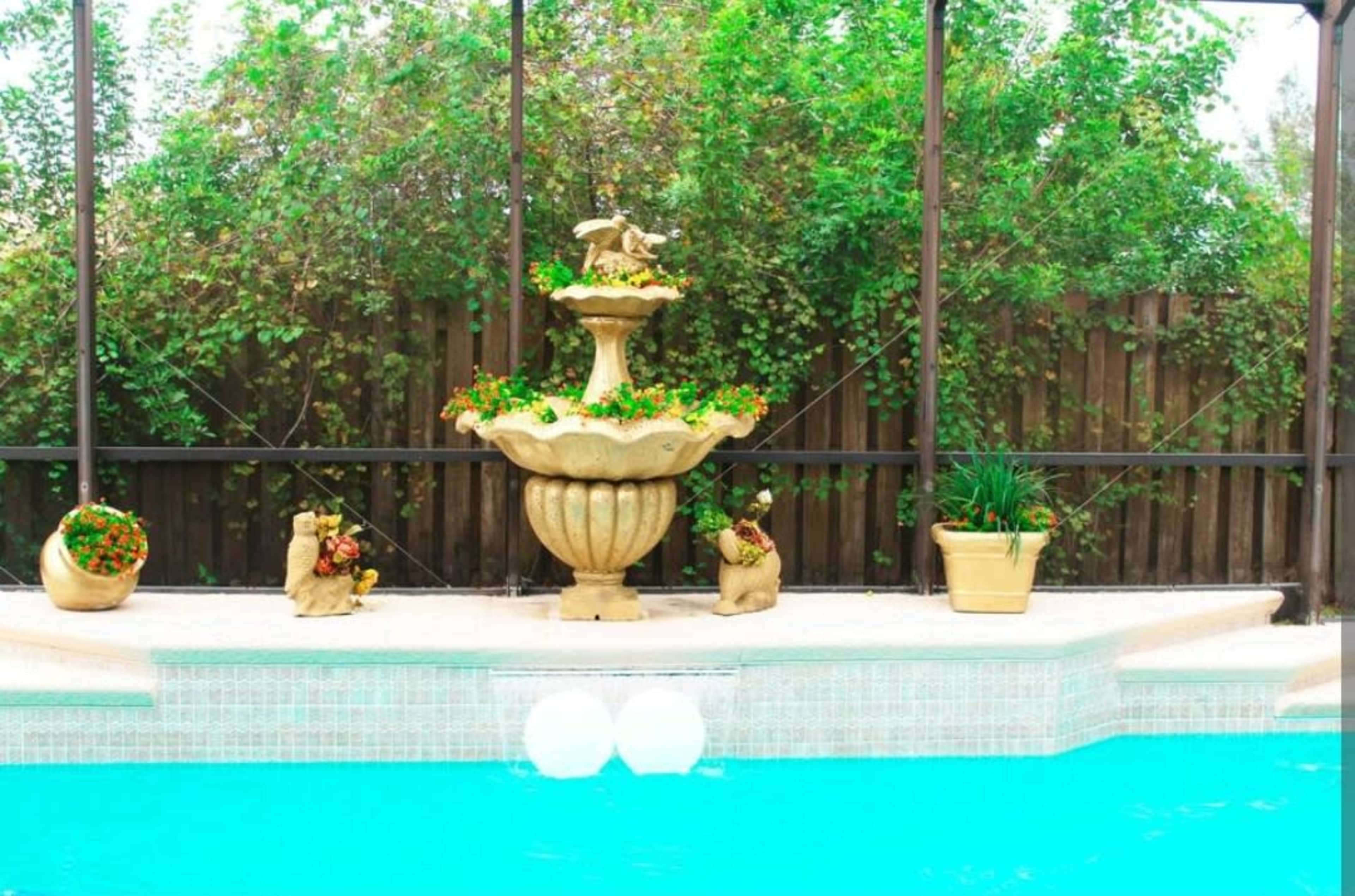 A decorative fountain sits amid potted plants beside a turquoise swimming pool, enclosed by a wire mesh structure.