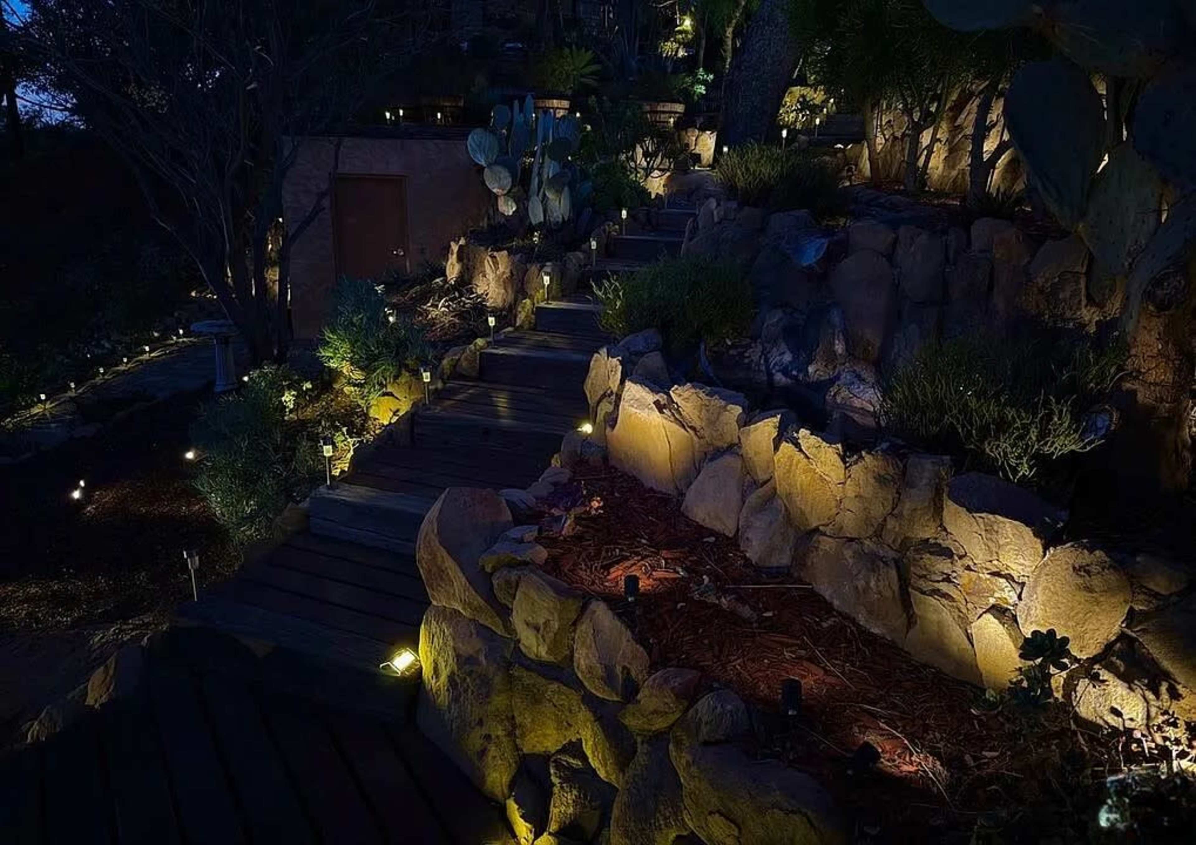 The image shows a landscaped pathway winding down a rocky slope, illuminated by soft outdoor lighting at night.