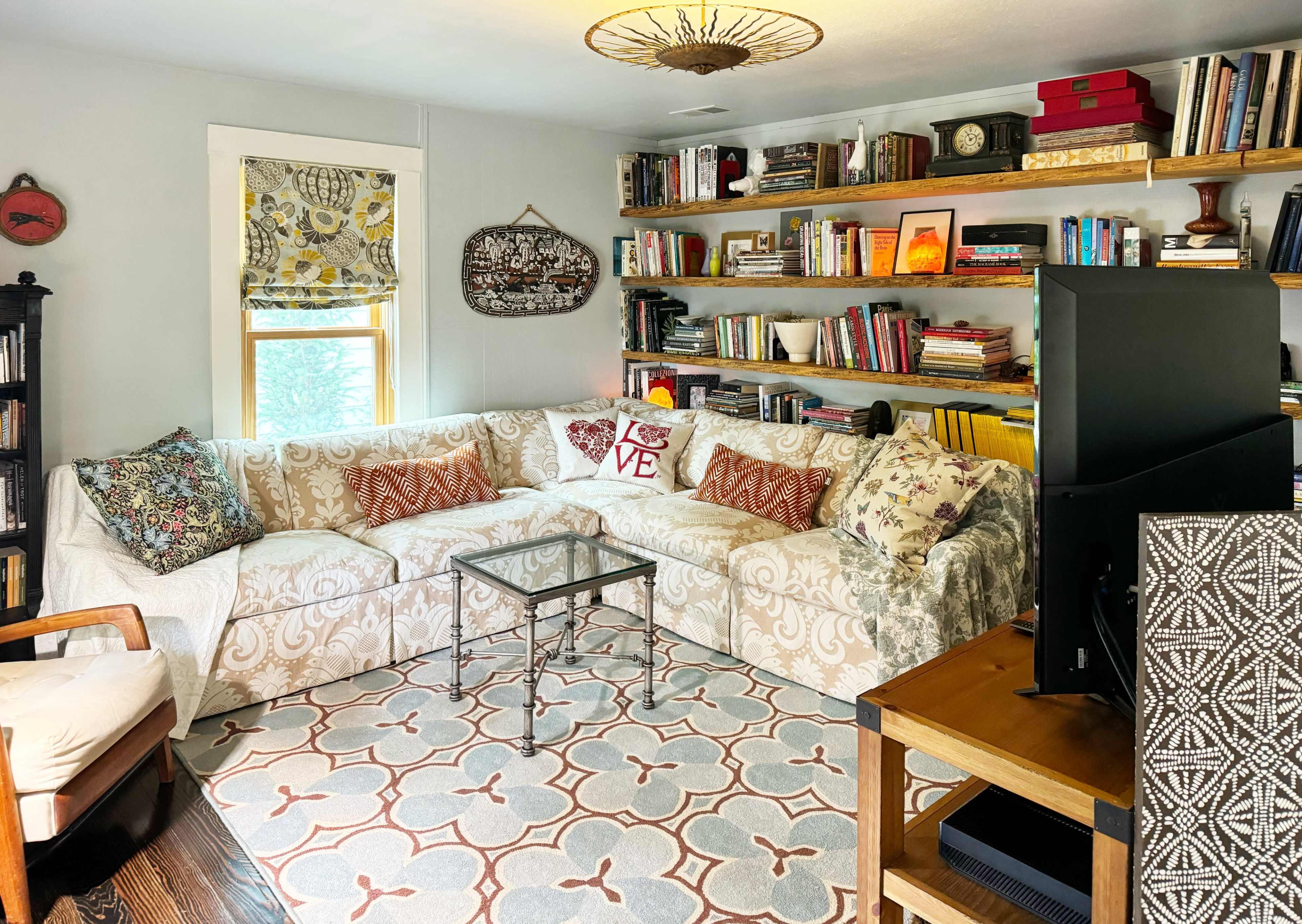 The image shows a cozy living room with a sectional sofa, a glass coffee table, shelves filled with books, and a television.