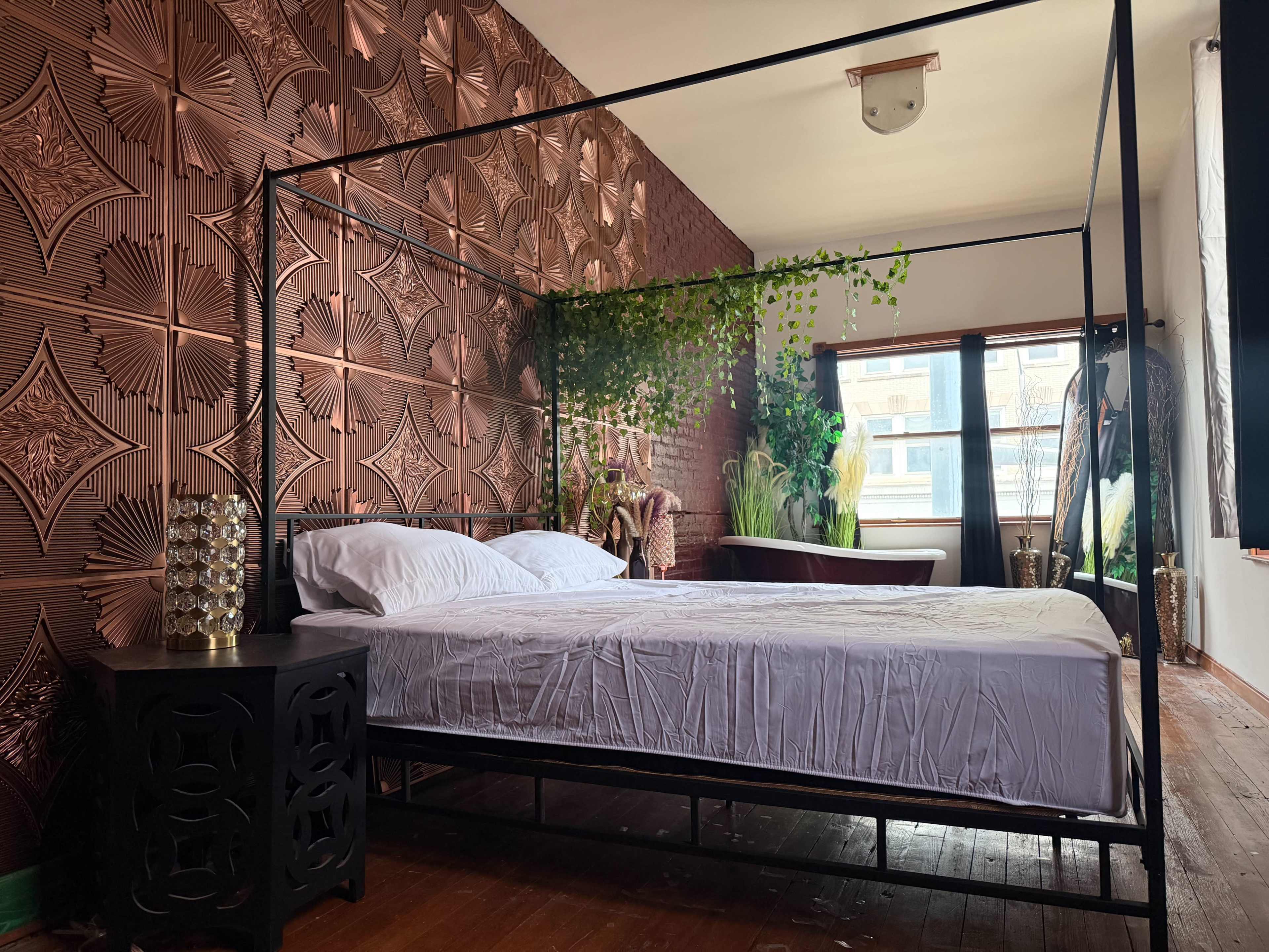 The image shows a bedroom with a framed bed, decorative wall panels, and various plants placed around the room.