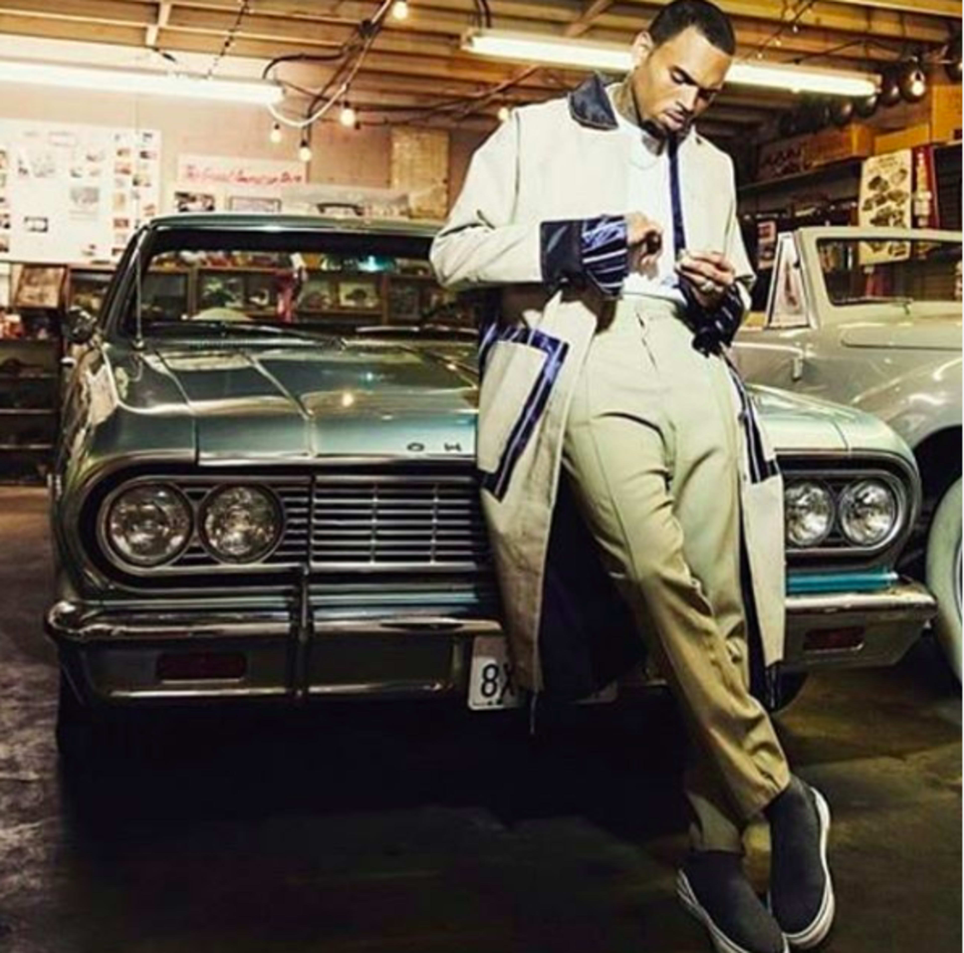 A man in a long coat stands beside a vintage car, checking his phone in a dimly lit garage filled with classic automobiles.