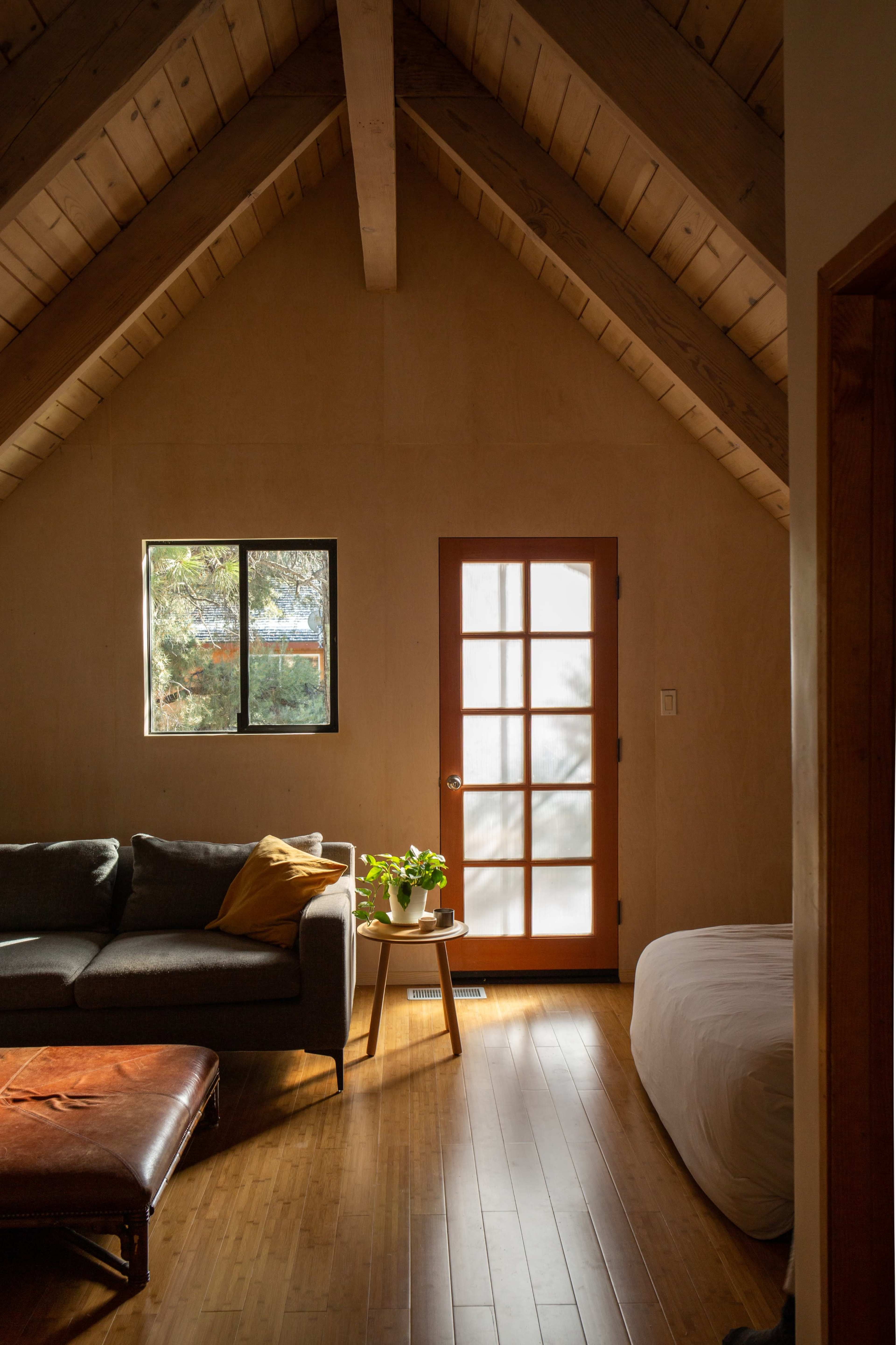 A cozy room with a sloped wooden ceiling, a grey sofa, a light brown leather ottoman, and a window illuminating a potted plant near a partially frosted door.